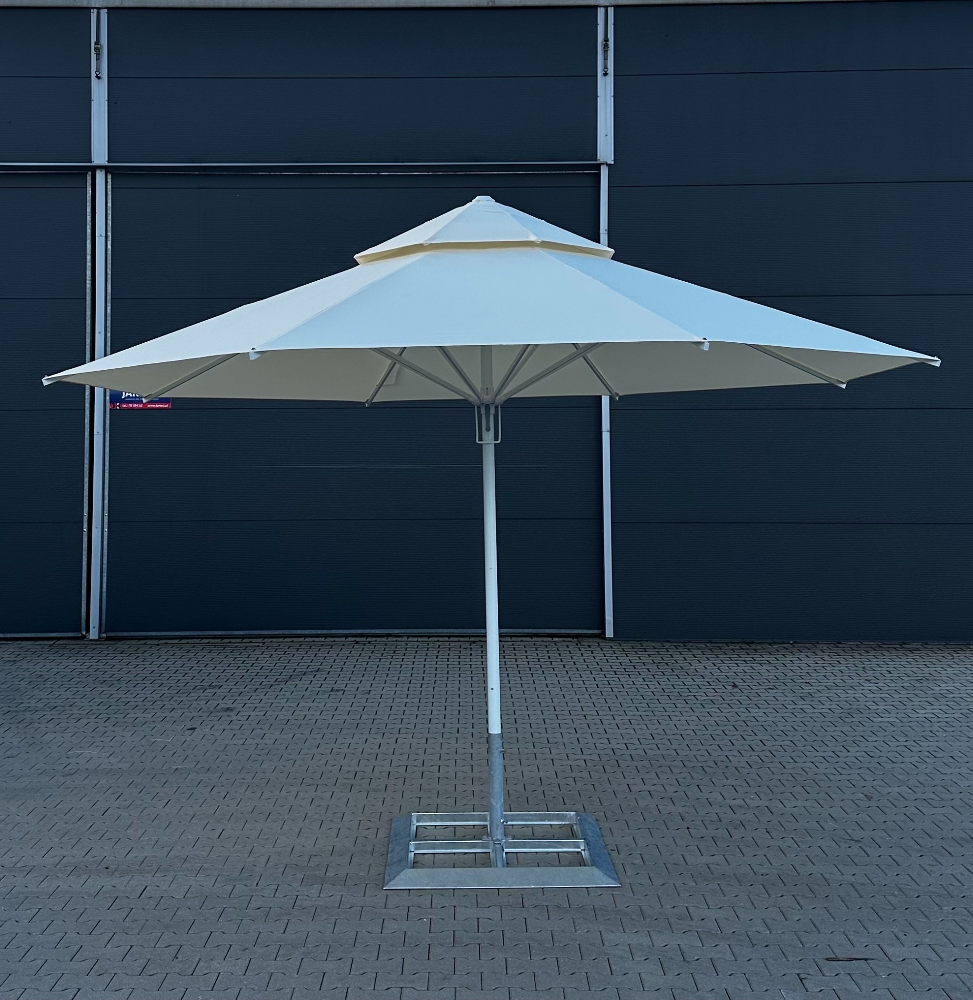 Rozłożony biały namiot parasolowy z dwupoziomowym dachem na metalowej podstawie, na tle szarej ściany hali i kostki brukowej.