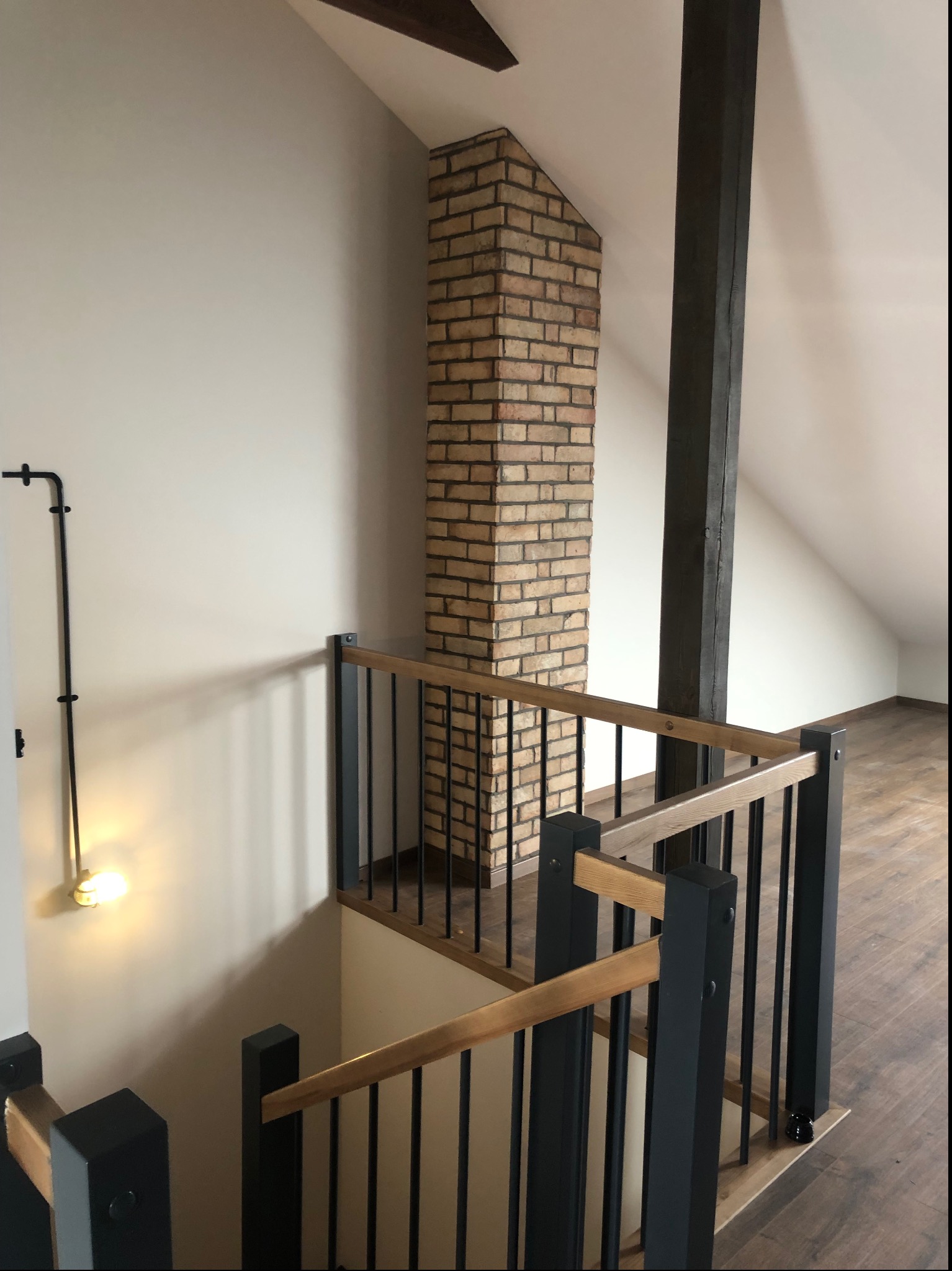 Poddasze z drewnianą podłogą, ceglanym kominem i czarno-drewnianą balustradą schodów, oświetlone kinkietem w industrialnym stylu.