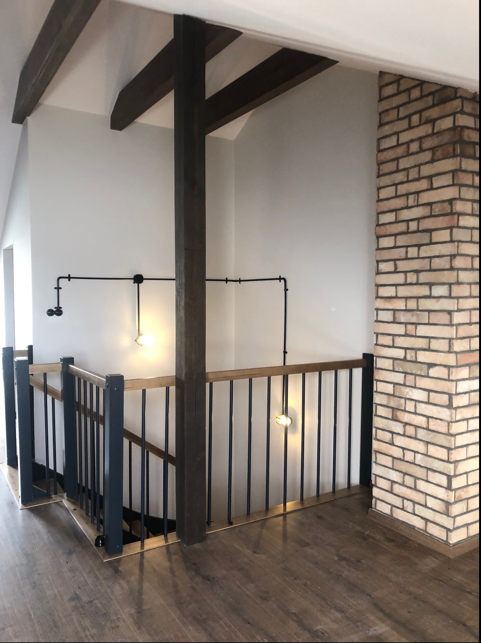 Nowoczesne wnętrze z drewnianymi belkami stropowymi, ceglanym filarem, czarną balustradą schodów i oświetleniem w stylu industrialnym.