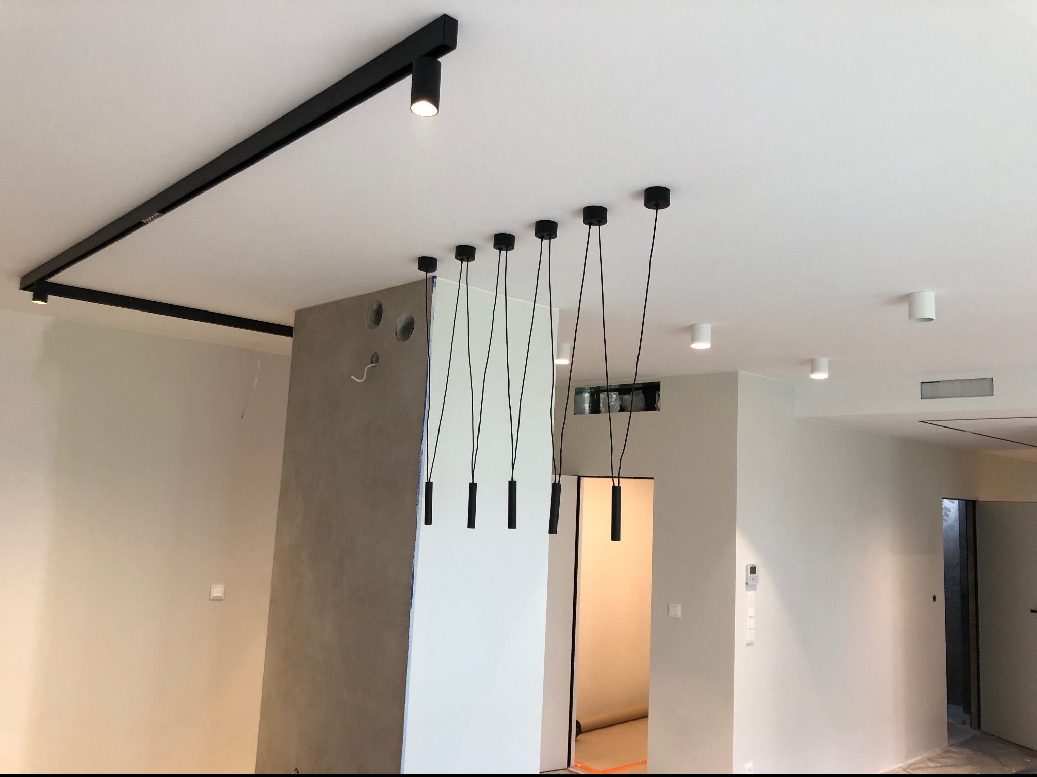 Nowoczesne oświetlenie sufitowe w stylu industrialnym w mieszkaniu w Warszawie: czarne szyny z reflektorami i wiszące lampy cylindryczne na tle białych ścian i sufitu.