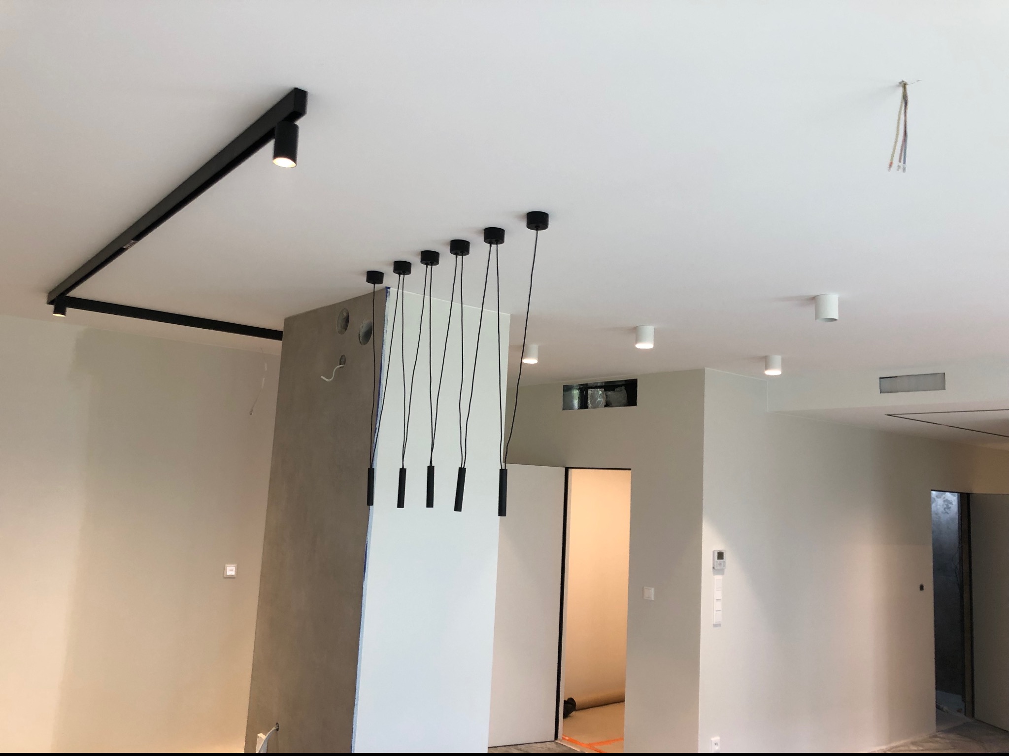 Nowoczesne oświetlenie sufitowe: czarne szyny z reflektorami punktowymi i wiszące lampy z minimalistycznymi, cylindrycznymi kloszami, na tle jasnego, odświeżonego wnętrza z elementami szarości...