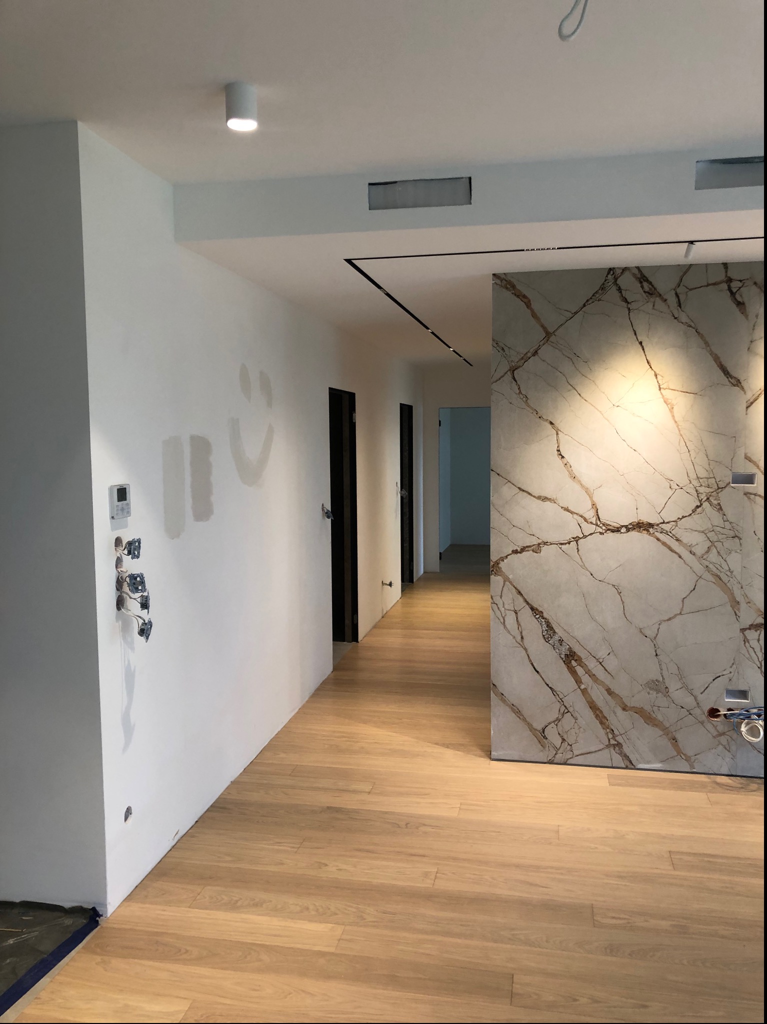 Korytarz w trakcie wykańczania: drewniana podłoga, ściana z imitacją marmuru, instalacje elektryczne w trakcie montażu, ślady po malowaniu na ścianie.