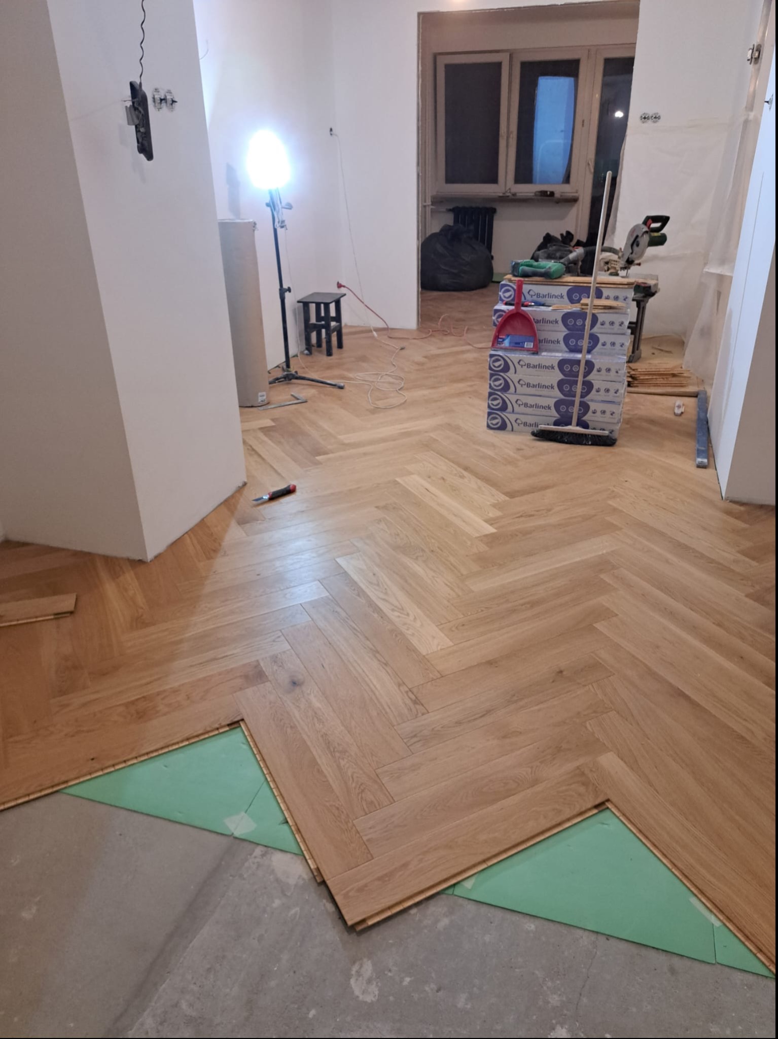 Układanie parkietu w jodełkę: widoczny fragment podkładu izolacyjnego, paczki paneli Barlinek, narzędzia i lampa budowlana w tle.