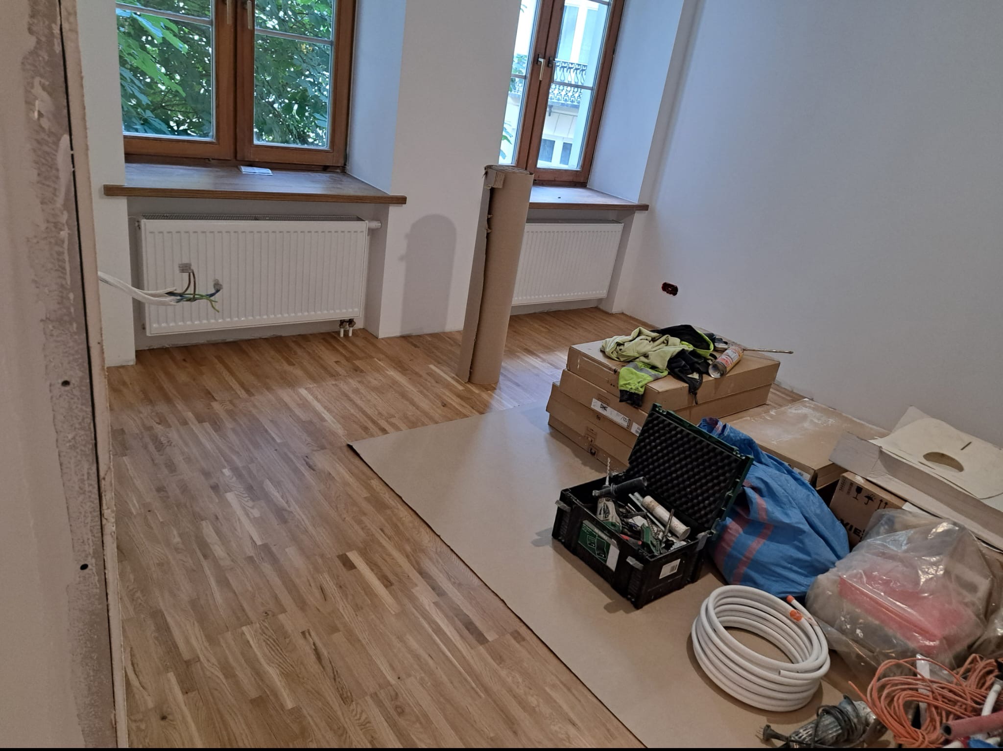 Pokój w trakcie prac wykończeniowych: drewniana podłoga zabezpieczona kartonem, narzędzia w kufrze, zwój białych rur, okna z drewnianą stolarką i grzejniki.