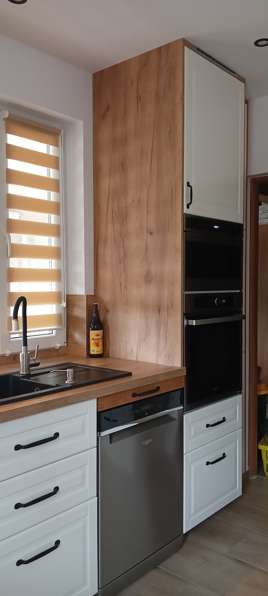 Nowoczesna kuchnia z białymi szafkami z czarnymi uchwytami, drewnianym blatem, czarnym zlewem i baterią, zabudowaną zmywarką oraz piekarnikiem w zabudowie. Na blacie stoi butelka piwa.