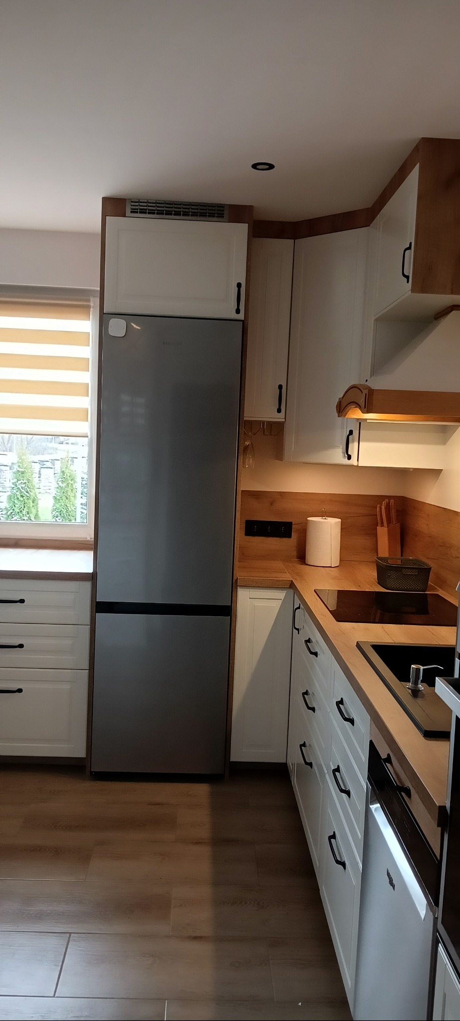 Nowoczesna kuchnia z białymi szafkami, drewnianym blatem i srebrną lodówką Samsung, widok na okno z żaluzjami poziomymi.