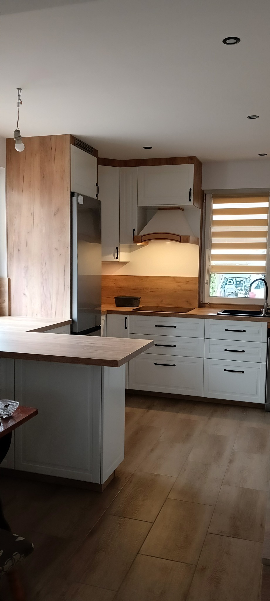 Jasna kuchnia z białymi szafkami i drewnianymi blatami, widok na okno z żaluzjami poziomymi i fragment podłogi z paneli imitujących drewno.