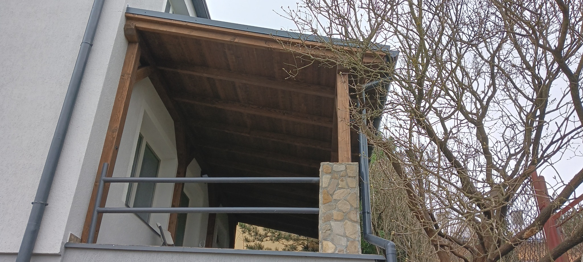 Zadaszony taras z drewnianą konstrukcją, filarem obłożonym kamieniem i metalową barierką, widok z zewnątrz na tle drzewa bez liści.