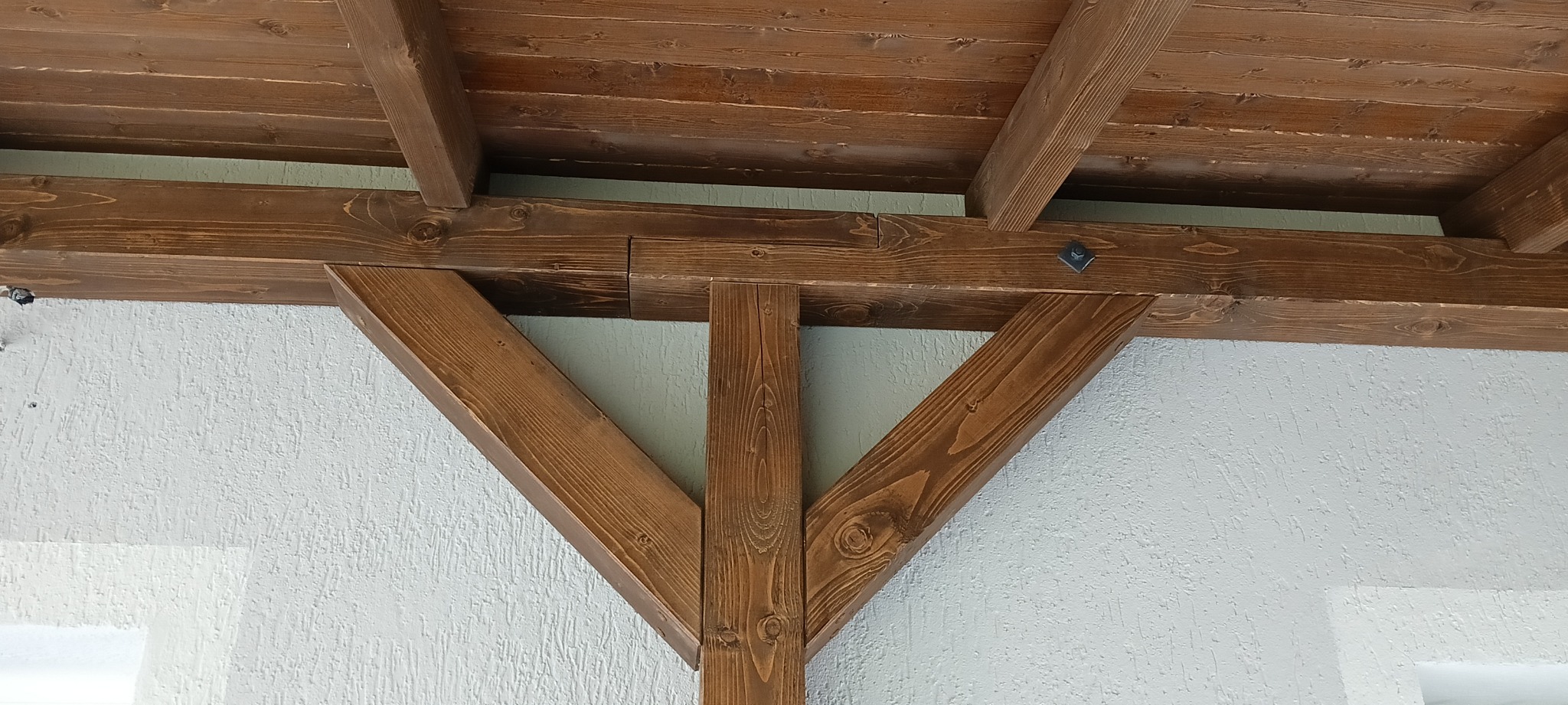 Drewniana konstrukcja z belek w kolorze ciemnego brązu, tworząca zadaszenie lub element dekoracyjny, przymocowana do białej, teksturowanej ściany.