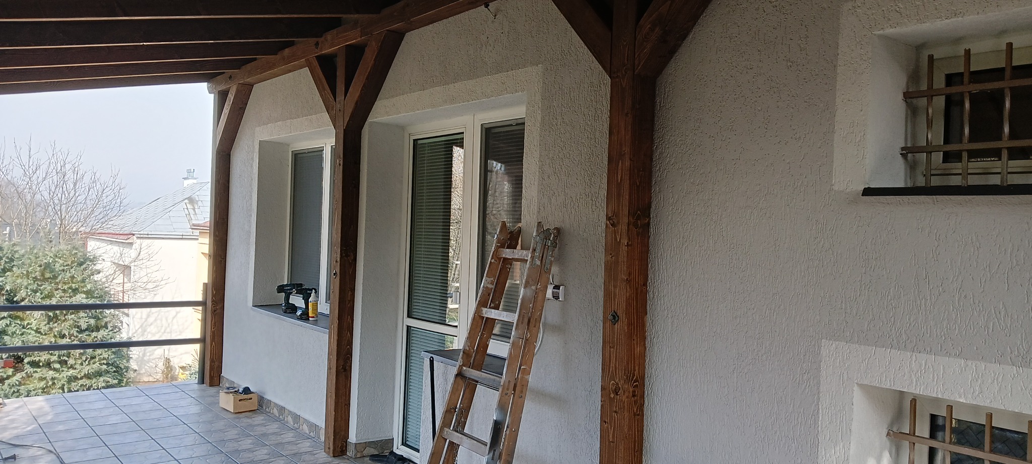 Zadaszony balkon z drewnianą konstrukcją nośną, świeżo otynkowane ściany zewnętrzne z oknami, drabina oparta o ścianę, narzędzia na parapecie okiennym.
