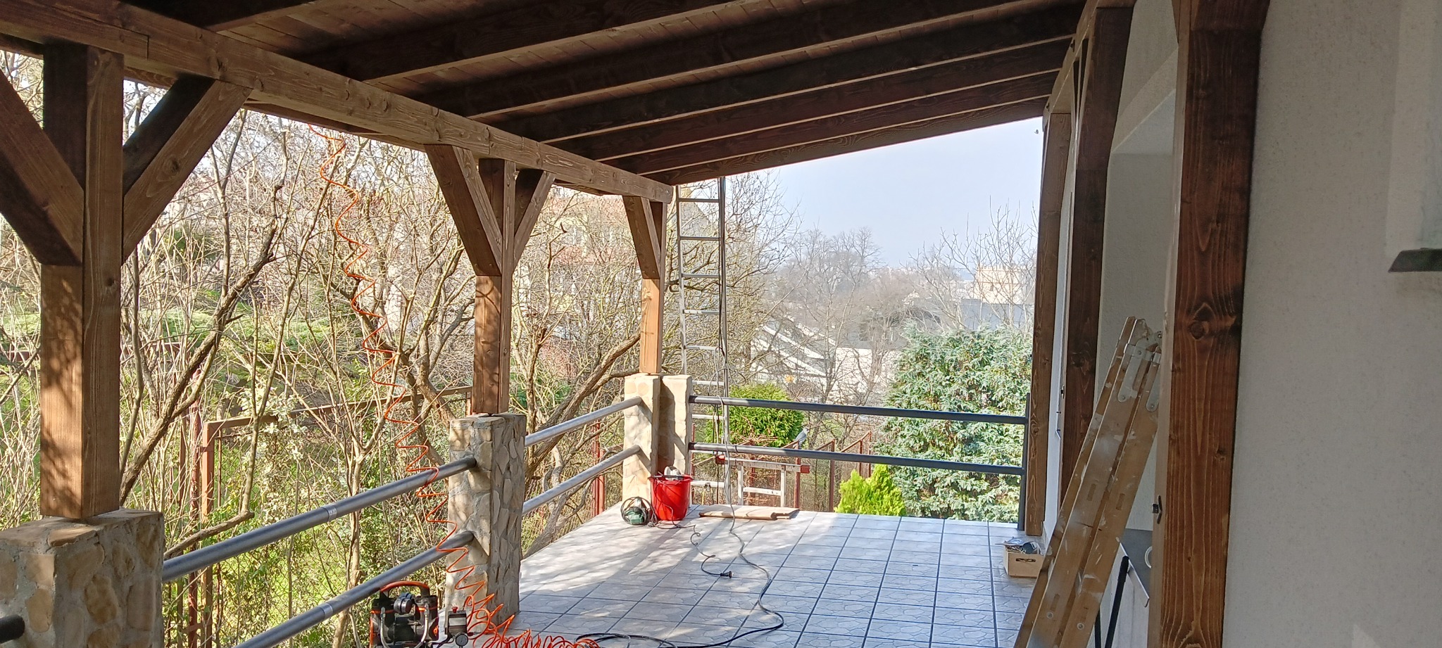 Zadaszony taras z drewnianą konstrukcją, balustradą z metalowych rurek i kamiennymi słupkami, widok na ogród, narzędzia pracy na podłodze
