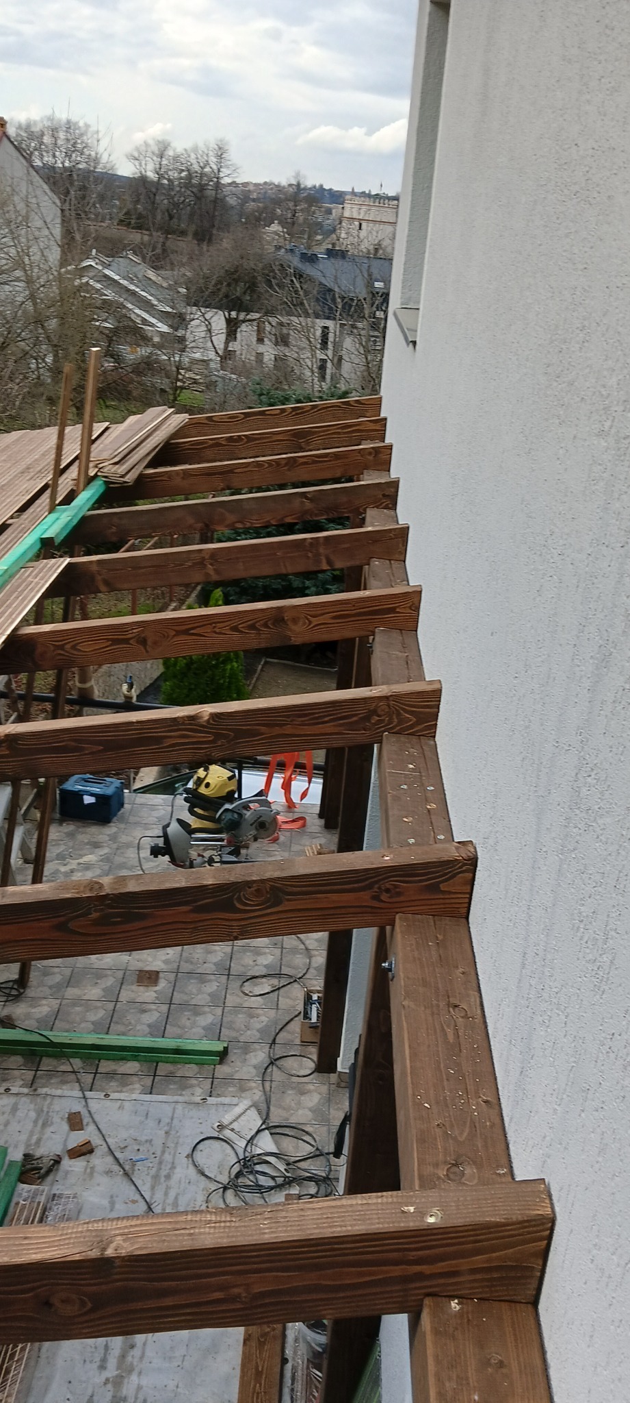 Budowa drewnianej pergoli przy ścianie budynku, widoczna ukośnica, deski i narzędzia, w tle zabudowa miejska.