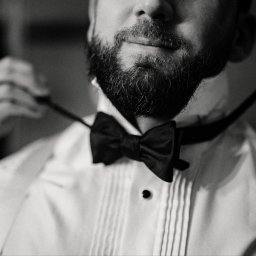 Ora Insieme - Czarno-białe zbliżenie brodatego mężczyzny w smokingu, poprawiającego muchę. Elegancki strój, przygotowania do uroczystości, subtelny uśmiech.