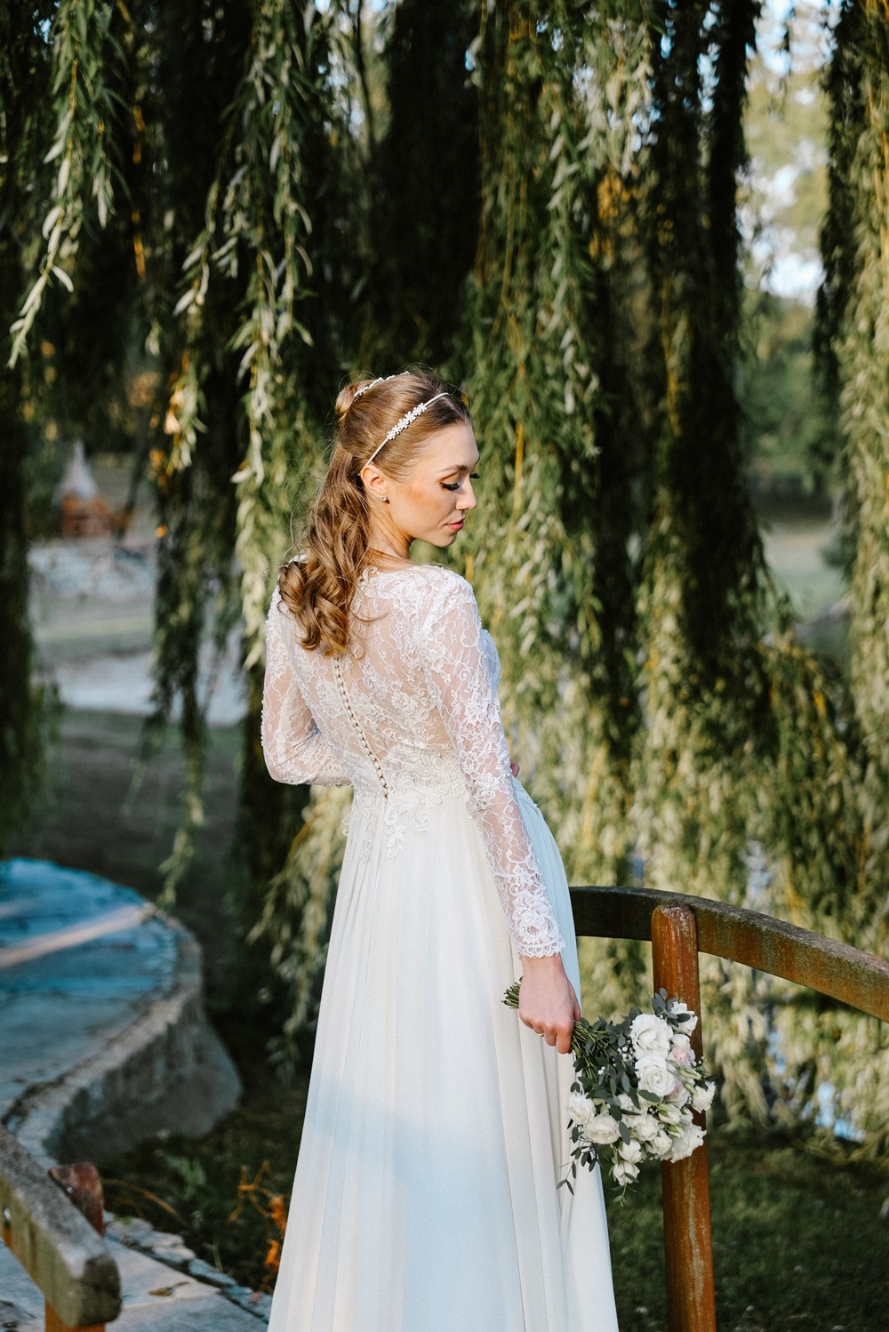 Panna młoda w koronkowej sukni, trzymająca bukiet, stoi na drewnianym mostku w otoczeniu zieleni. Widok z tyłu, delikatne oświetlenie.