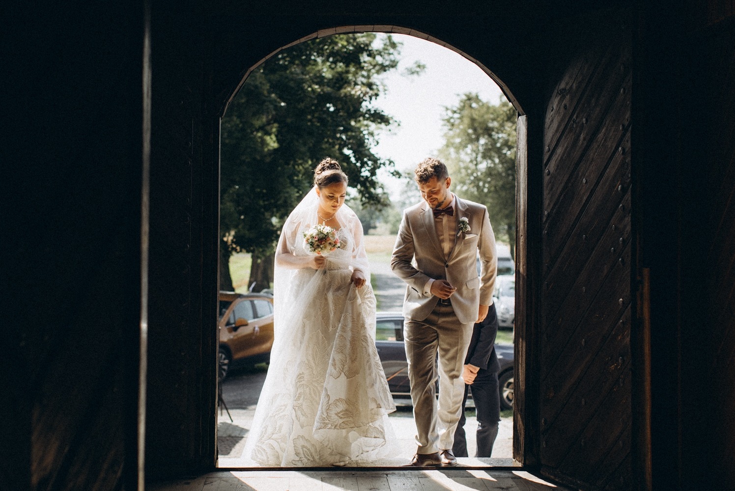 Młoda para wychodzi z kościoła, panna młoda w koronkowej sukni z bukietem, pan młody w beżowym garniturze z muchą, w tle zieleń drzew i zaparkowane samochody.
