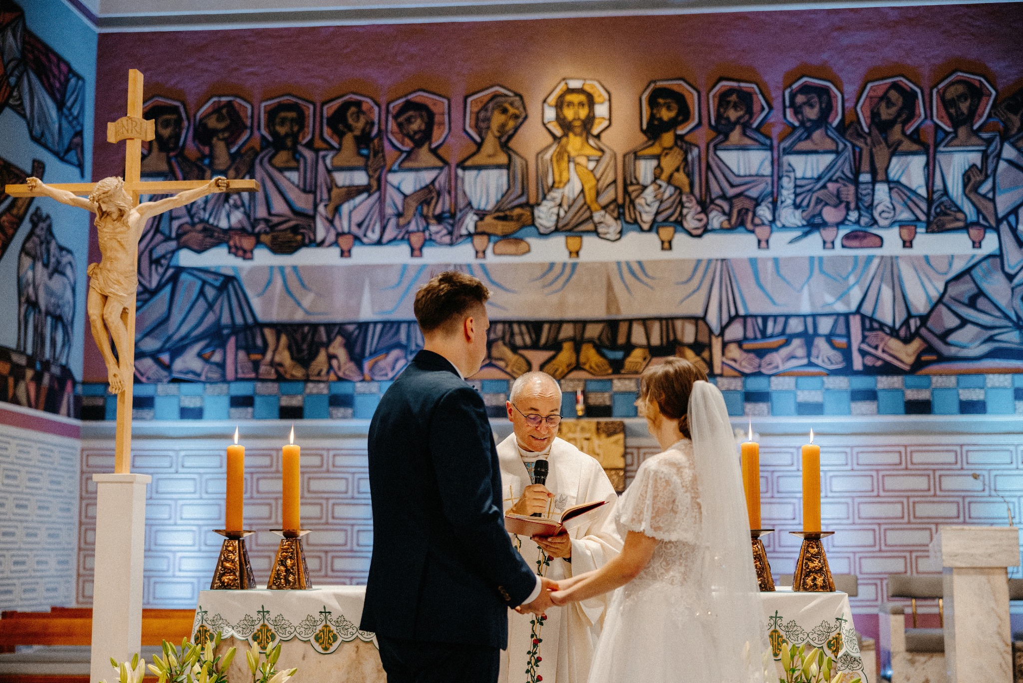 Uroczystość ślubna w kościele: para młoda trzyma się za ręce przed księdzem, w tle ołtarz z krzyżem i świecami, oraz malowidło Ostatniej Wieczerzy.