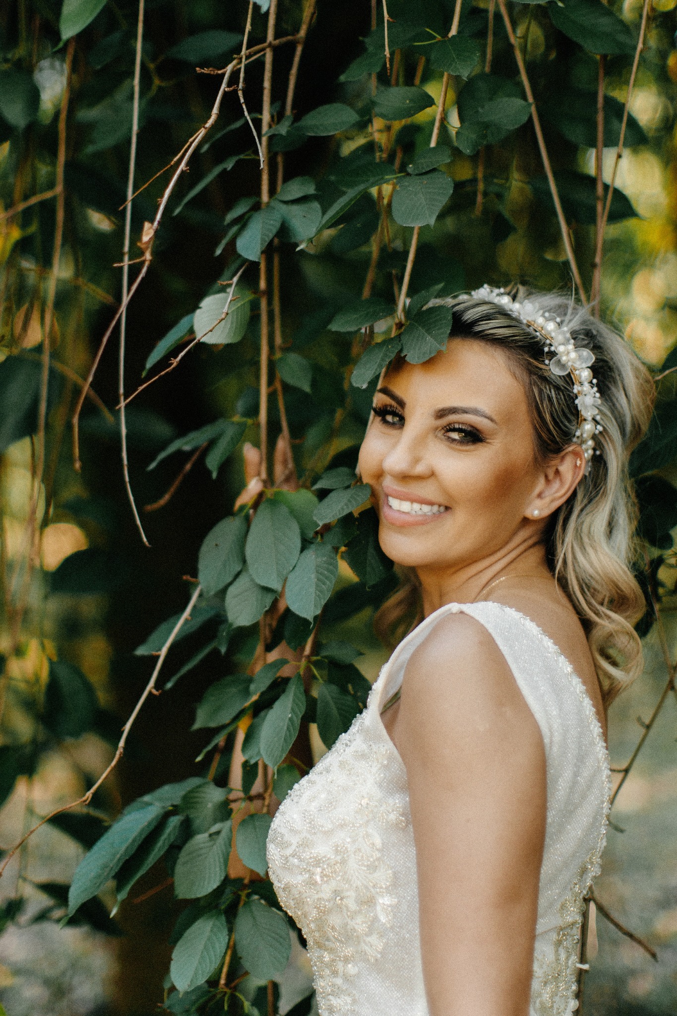 Uśmiechnięta panna młoda w białej sukni z perłowym diademem, pozująca na tle zielonych liści. Delikatne oświetlenie podkreśla makijaż i naturalny urok.