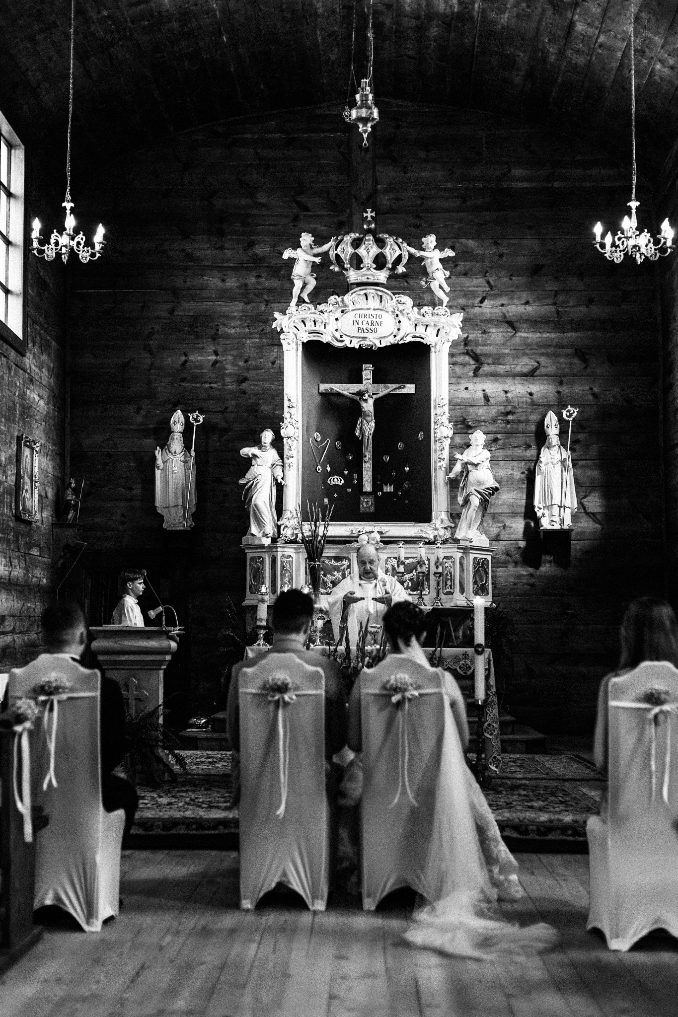 Czarno-białe zdjęcie wnętrza kościoła podczas ceremonii ślubnej. Widoczni państwo młodzi, ksiądz przy ołtarzu i goście siedzący na krzesłach z dekoracjami.