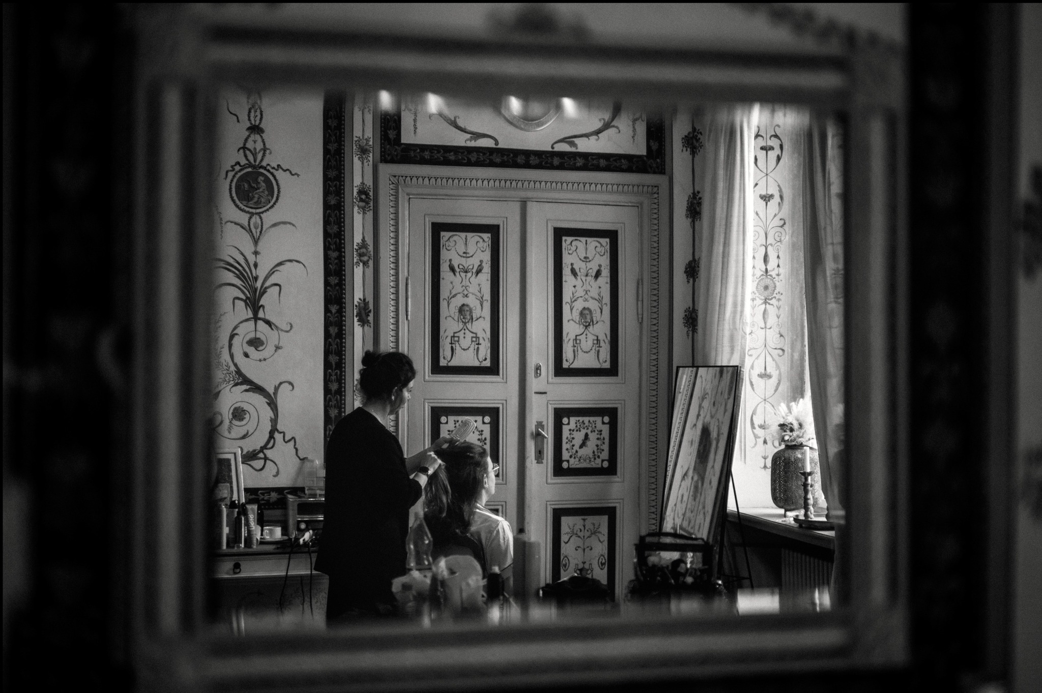 Czarno-białe ujęcie zza ramy lustra: fryzjerka czesze pannę młodą na tle bogato zdobionych drzwi i ścian w stylu retro, przygotowania do ślubu.