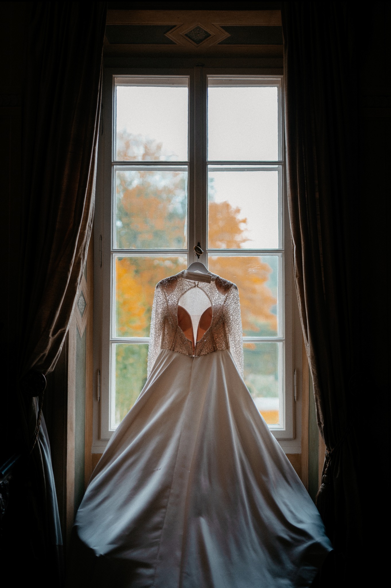 Elegancka suknia ślubna z koronkowymi rękawami, wisząca na tle okna z widokiem na jesienne drzewa. Subtelne światło podkreśla detale kreacji.