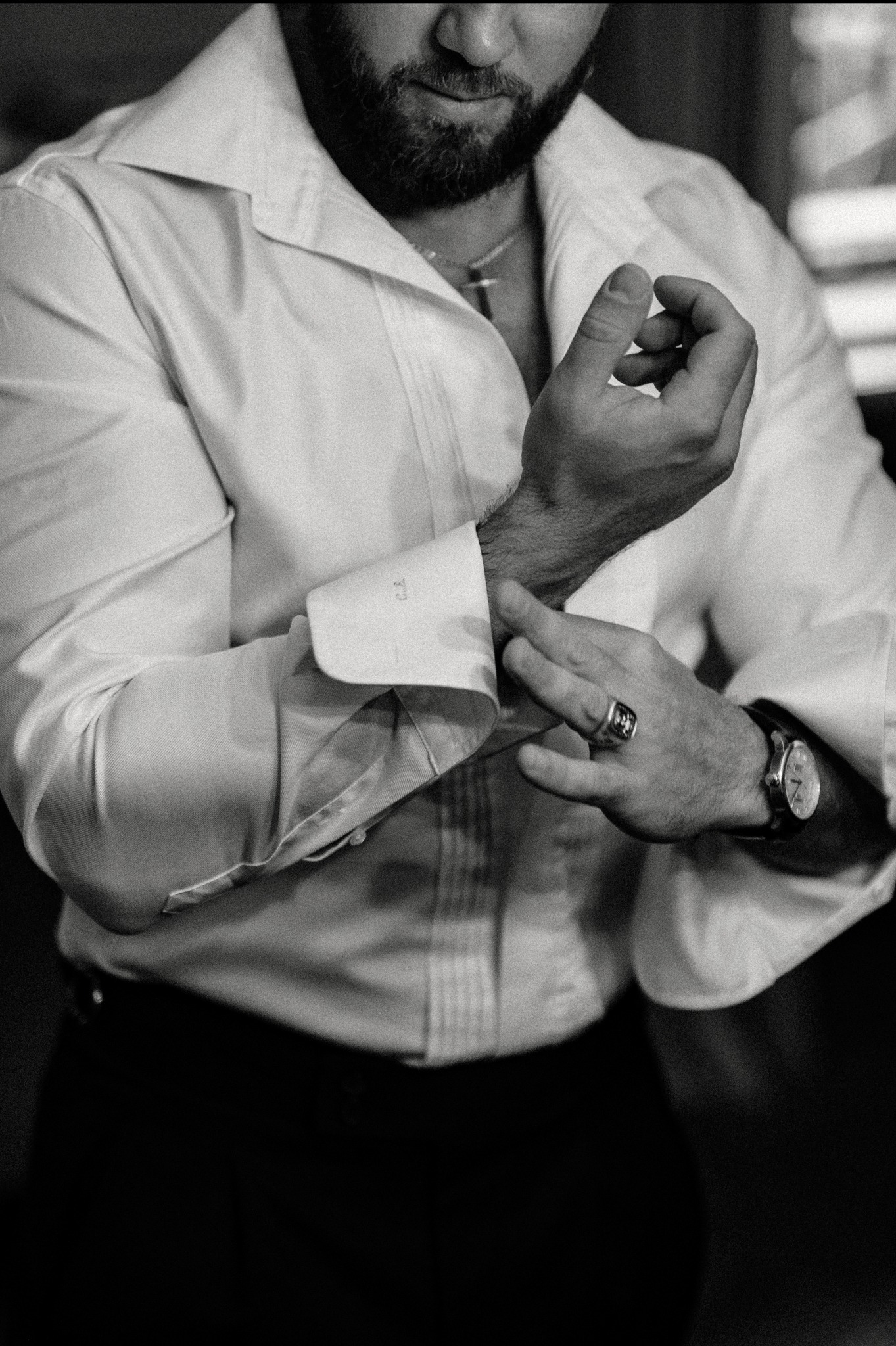 Czarno-białe zbliżenie na mężczyznę zapinającego mankiet koszuli z monogramem, widoczny zegarek i sygnet, przygotowania do uroczystości.