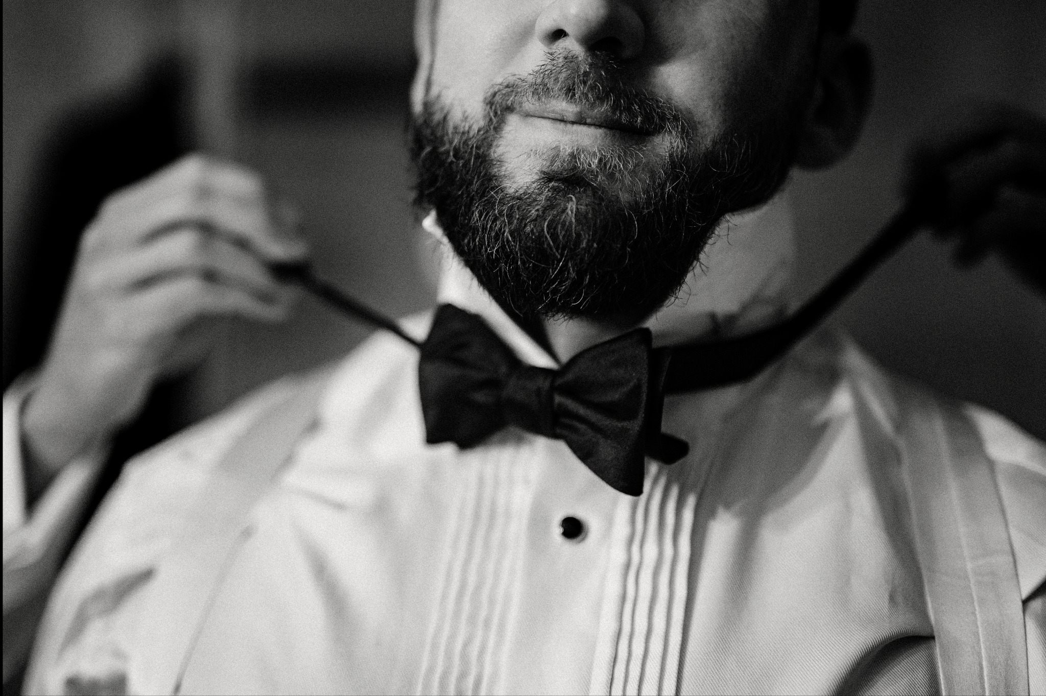 Czarno-białe zbliżenie brodatego mężczyzny w smokingu, poprawiającego muchę. Elegancki strój, przygotowania do uroczystości, subtelny uśmiech.