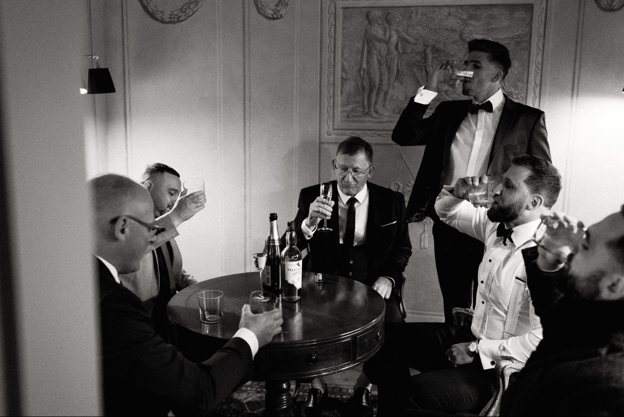 Czarno-białe zdjęcie grupy elegancko ubranych mężczyzn wznoszących toast w pomieszczeniu z dekoracyjnym obrazem na ścianie. Ujęcie z dołu, podkreślające uroczysty charakter.