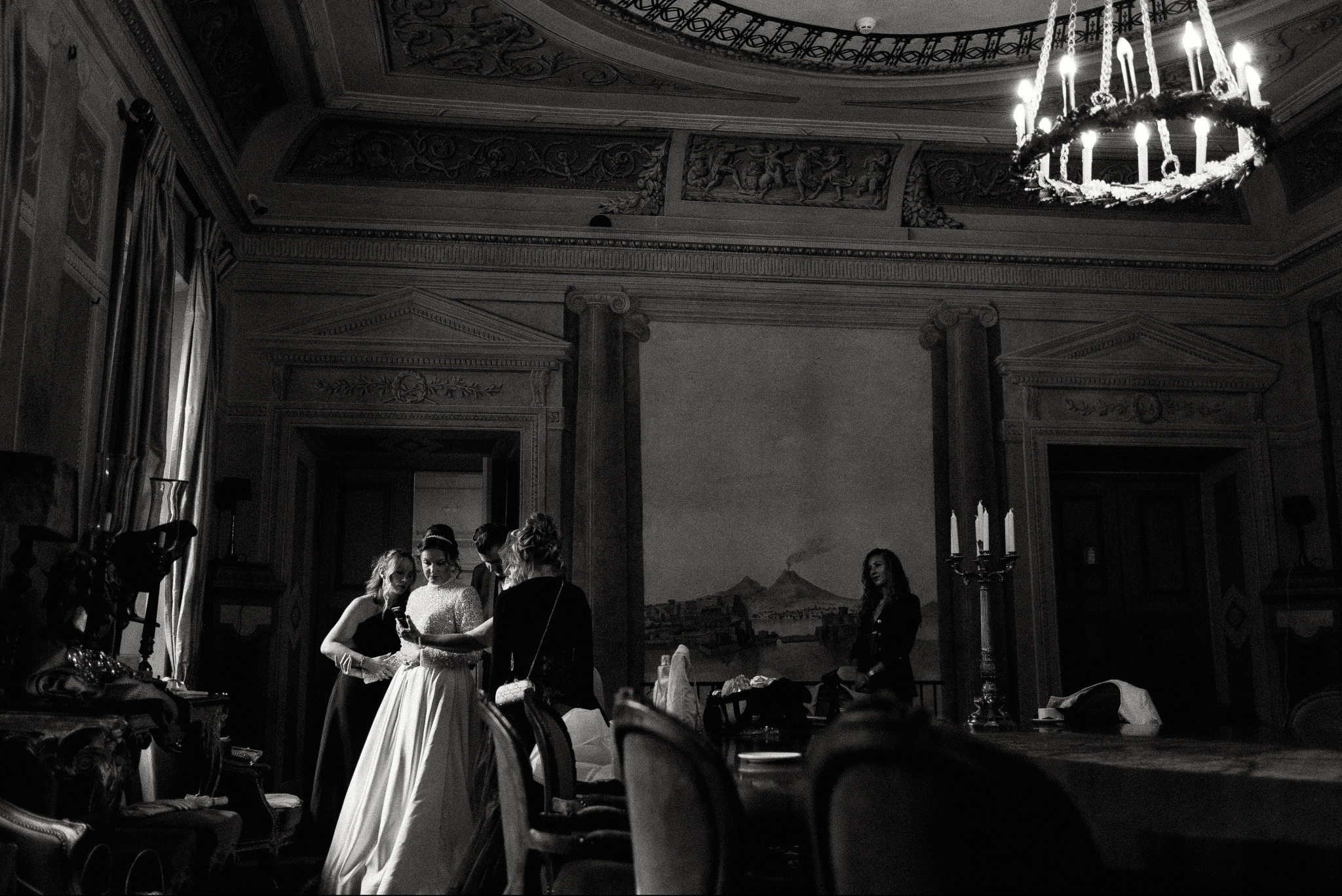 Czarno-białe zdjęcie panny młodej w bogato zdobionym wnętrzu pałacowym, otoczonej przez druhny. W tle widoczny żyrandol i malowidła ścienne z motywem wulkanu.