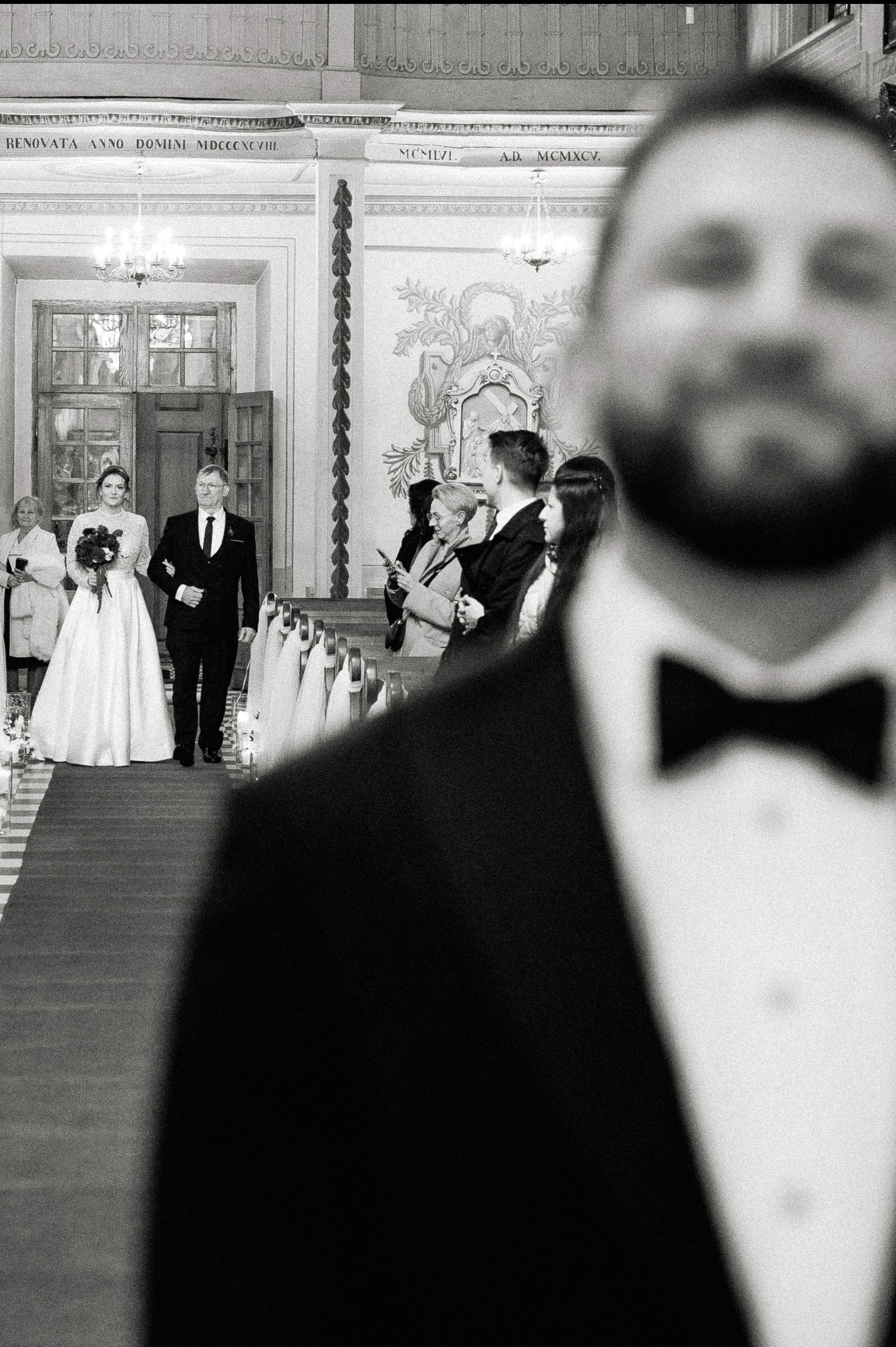 Czarno-białe zdjęcie z kościoła: panna młoda z ojcem idą do ołtarza, w tle goście, na pierwszym planie zamazana sylwetka mężczyzny w muszce.