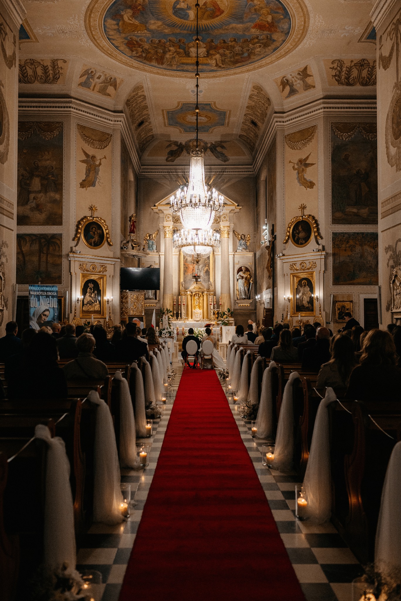 Para młoda siedzi przed ołtarzem w bogato zdobionym kościele. Czerwony dywan, świece i dekoracje na ławkach tworzą uroczystą atmosferę.