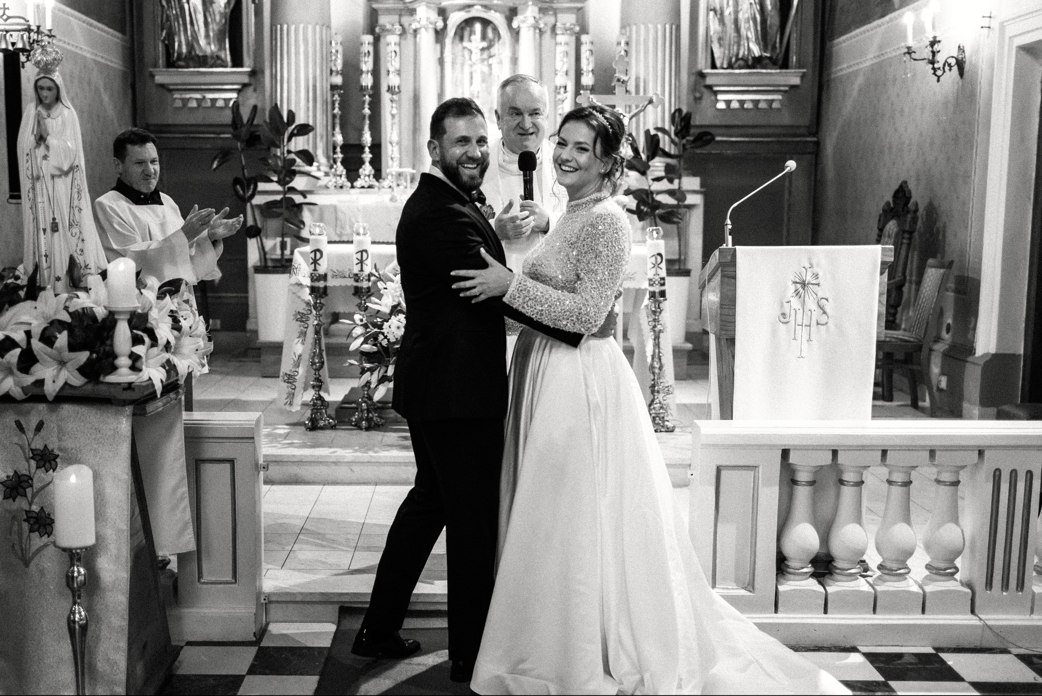 Czarno-białe zdjęcie uśmiechniętej pary młodej w kościele podczas ceremonii ślubnej, z księdzem w tle i dekoracjami kwiatowymi.