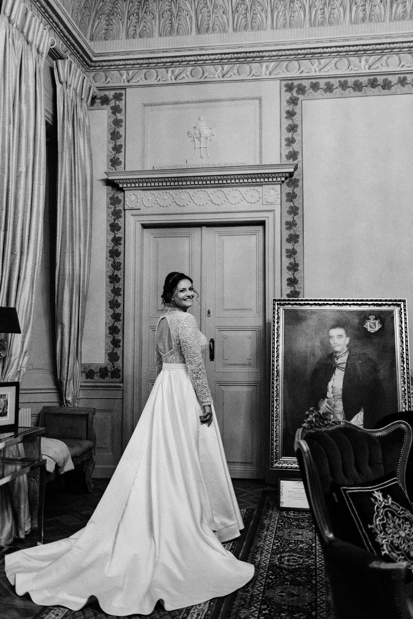Panna młoda w sukni z odkrytymi plecami, spogląda przez ramię w bogato zdobionym wnętrzu pałacowym z portretem w tle. Czarno-biała fotografia ślubna.