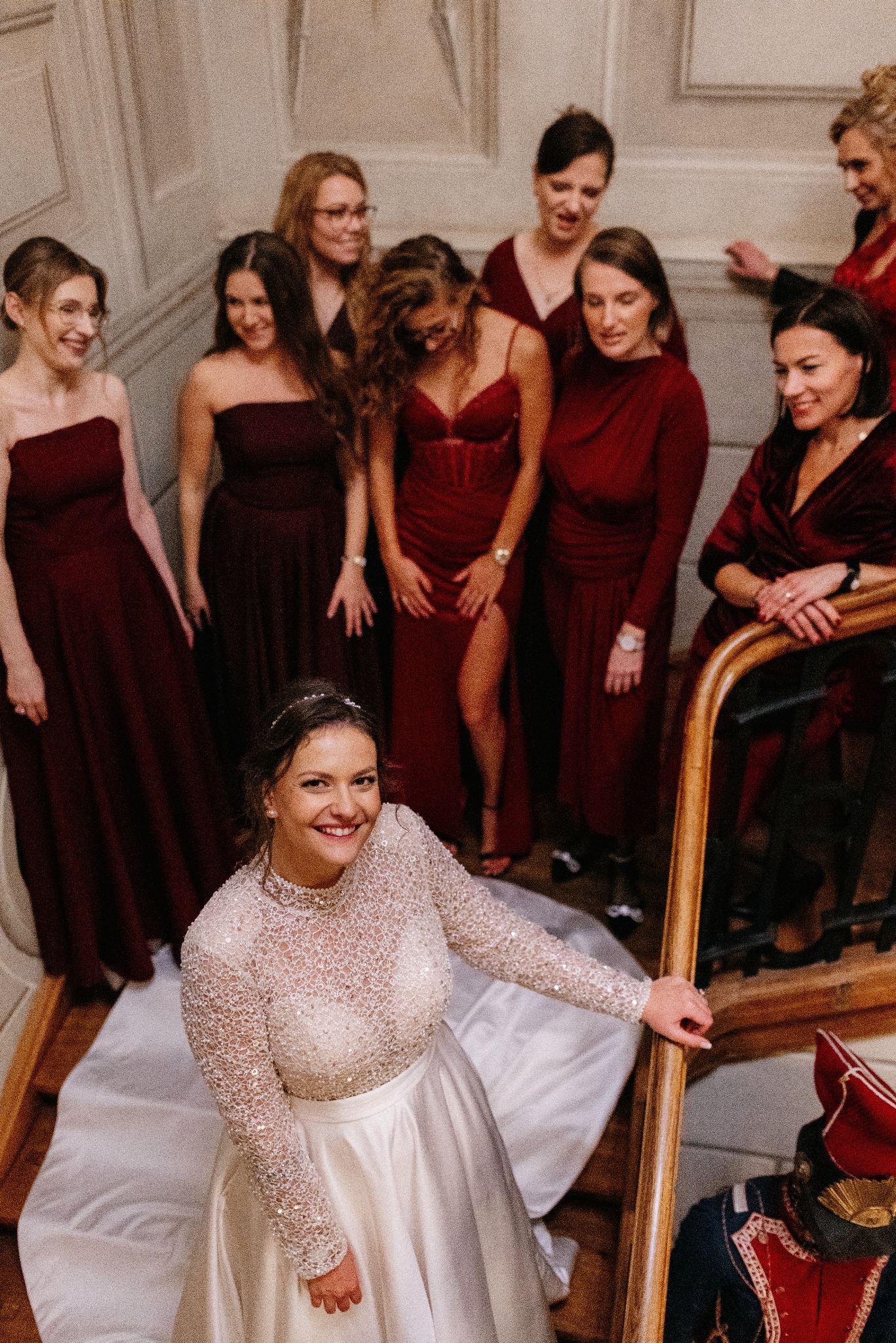 Uśmiechnięta panna młoda na schodach z druhnami w bordowych sukienkach. Nietypowy strój galowy w tle. Zdjęcie z góry, ciepłe oświetlenie.