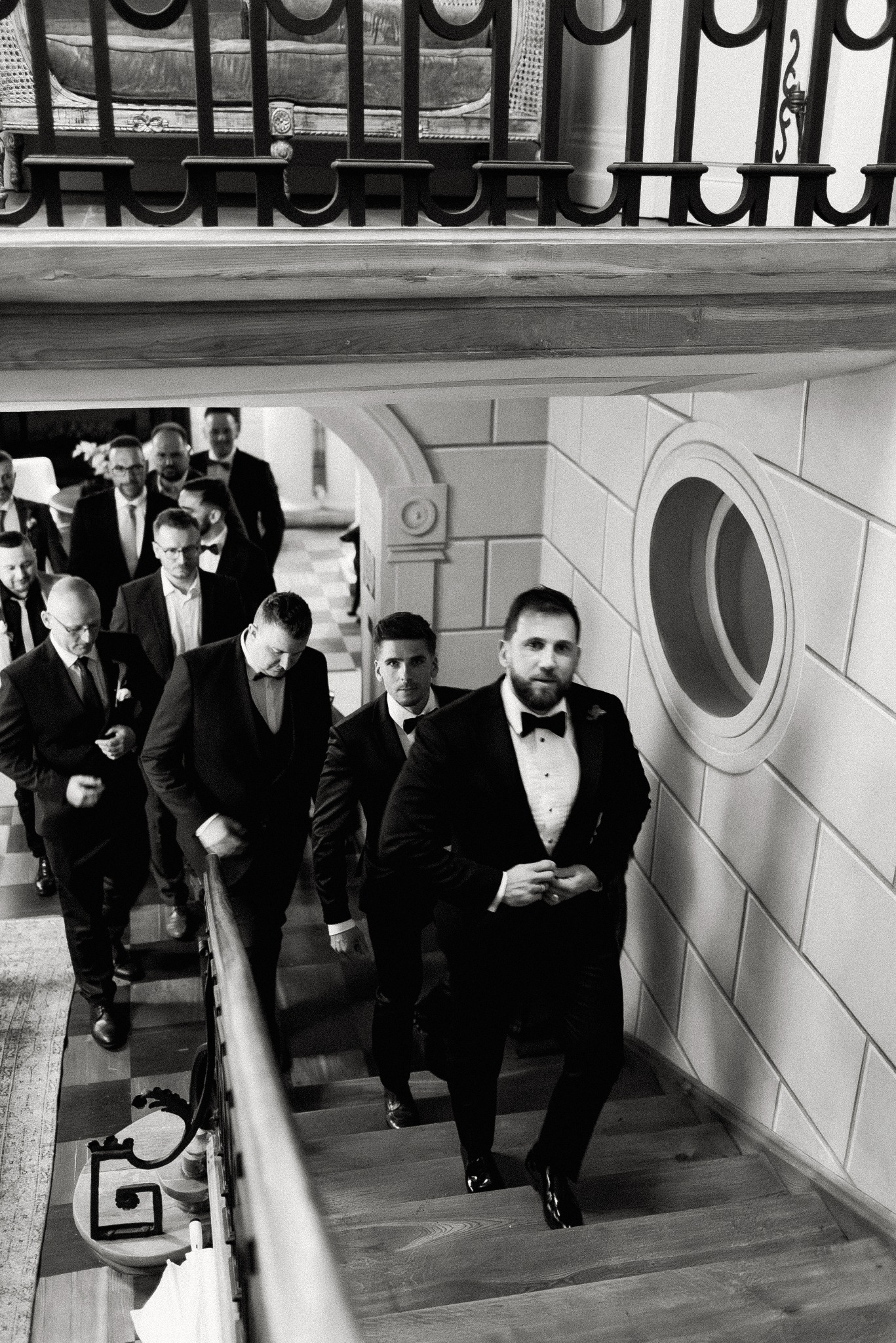Czarno-białe zdjęcie grupy elegancko ubranych mężczyzn w smokingach, wchodzących po schodach w budynku z dekoracyjnymi elementami architektonicznymi.