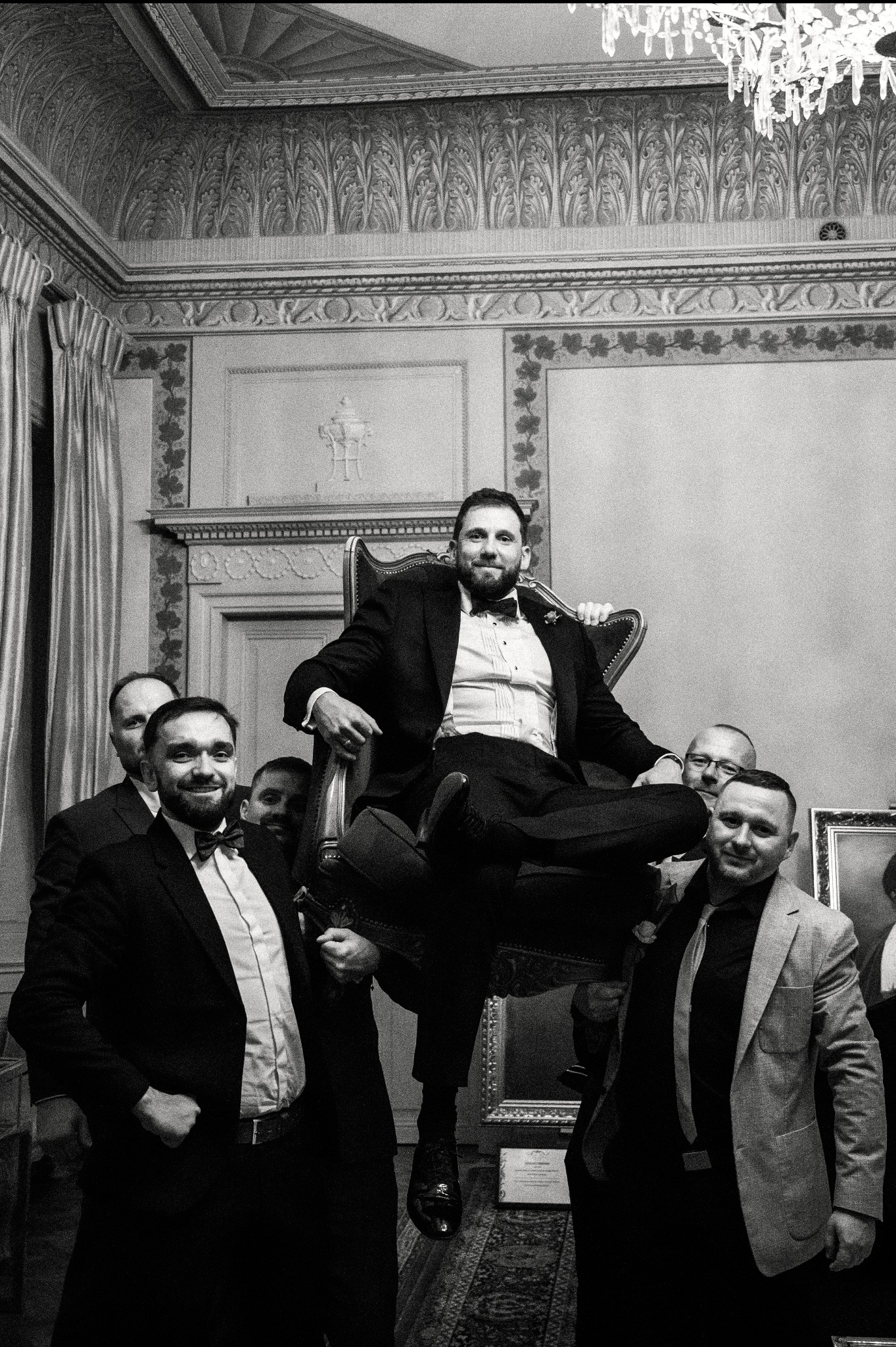 Czarno-białe zdjęcie: Uśmiechnięty pan młody w fotelu niesionym przez gości weselnych w bogato zdobionym wnętrzu. Eleganckie stroje, żyrandol, dekoracje ścian.