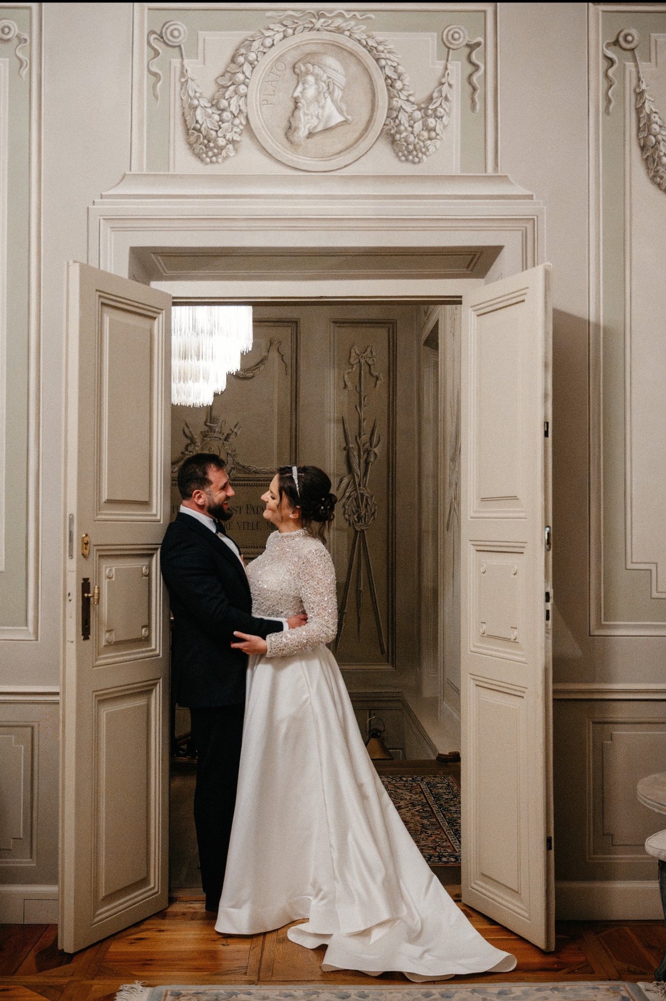 Para młoda w objęciach, w eleganckim wnętrzu z otwartymi drzwiami i dekoracyjnymi płaskorzeźbami, ciepłe oświetlenie, suknia z długim rękawem.