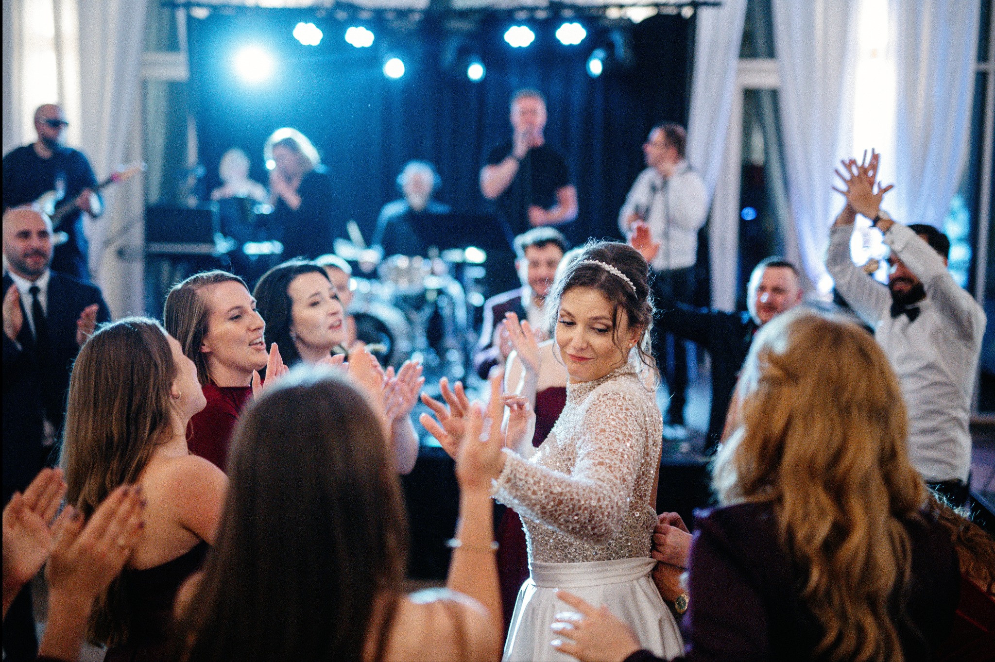 Panna młoda w błyszczącej sukni tańczy z gośćmi weselnymi na parkiecie, w tle zespół muzyczny i oświetlenie sceniczne.