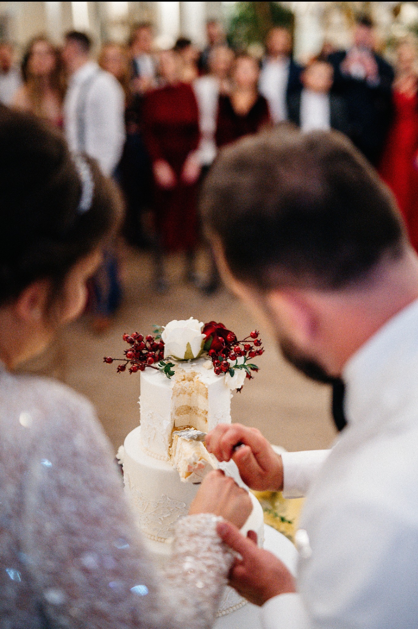 Para młoda kroi tort weselny, dekorowany kwiatami i owocami, na tle gości. Ujęcie zza pleców, skupione na torcie i dłoniach.