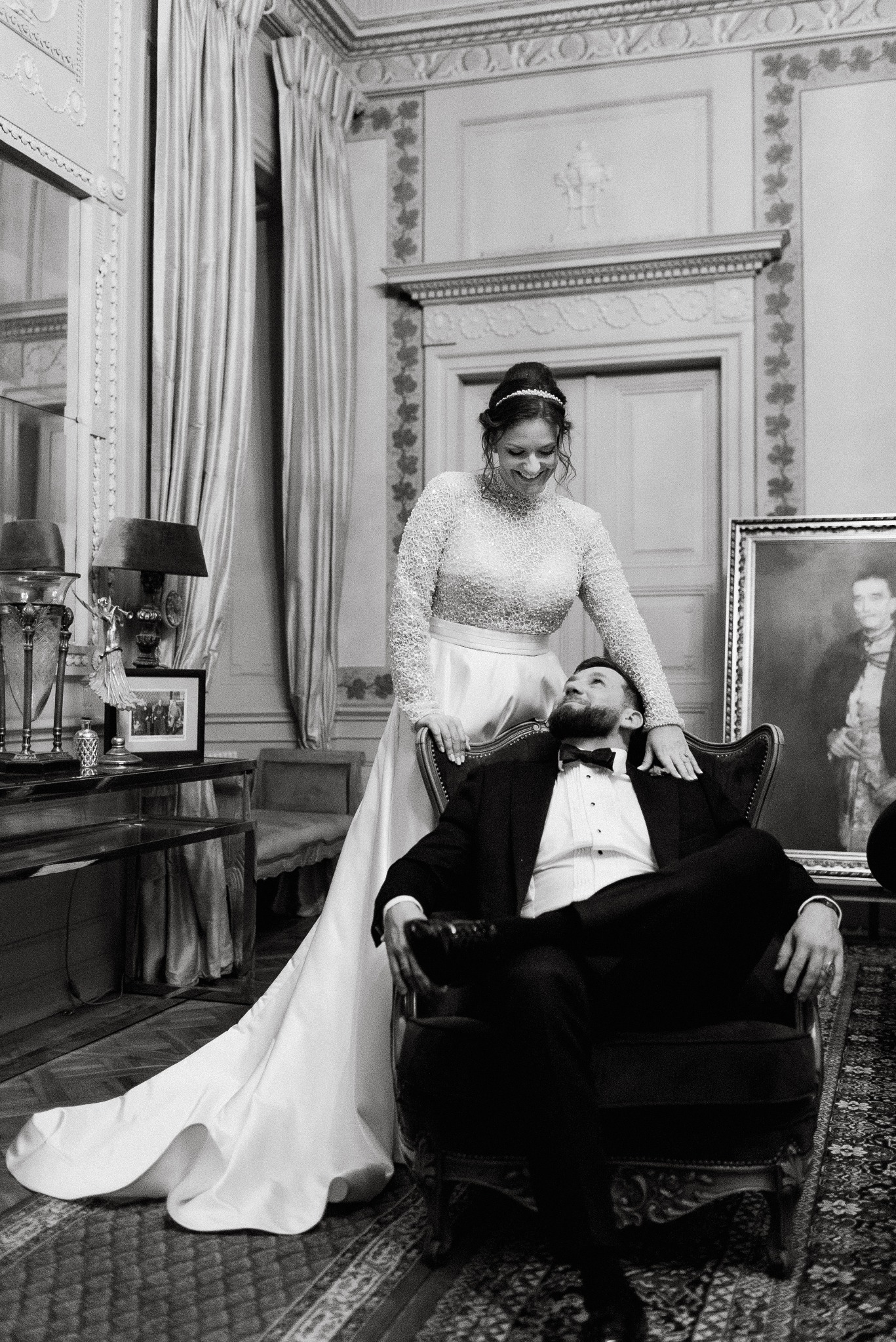 Czarno-białe zdjęcie pary: panna młoda w sukni z długim rękawem opiera się o fotel, w którym siedzi pan młody w smokingu, oboje patrzą na siebie z uśmiechem, w tle bogato zdobione wnętrze.