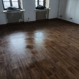 Fantazy - Nowo ułożona, ciemnobrązowa podłoga z paneli w pustym pokoju z dwoma oknami. Okna zabezpieczone folią. Na parapetach leżą worki z gruzem. Widoczne gniazdka elektryczne na ścianie.