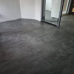 Fantazy - Szara podłoga w nowym pomieszczeniu z widocznymi kablami elektrycznymi wystającymi ze ściany, pracownik zamiata podłogę przed dalszymi pracami wykończeniowymi.