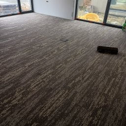 Fantazy - Wzorzysta, ciemna podłoga w nowym pomieszczeniu z dużymi oknami, widoczny fragment zwiniętego dywanu i żółta rura na zewnątrz.