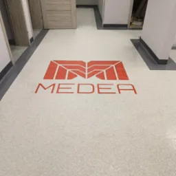 Jasnoszara podłoga w nowym budynku z czerwonym logo 'MEDEA' w centralnej części, widoczne drzwi i fragmenty korytarza.