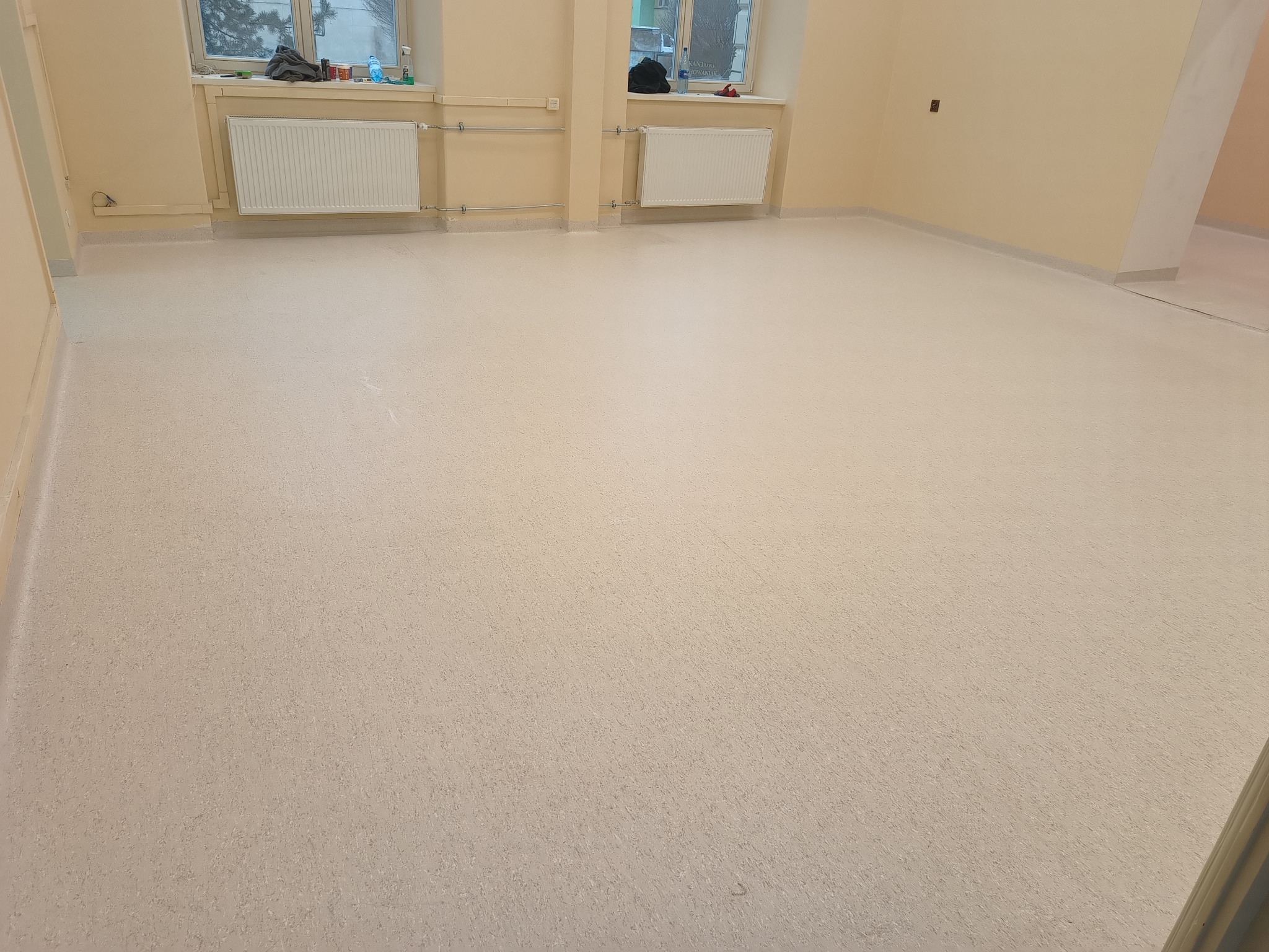 Jasne, puste pomieszczenie z nową, jednolitą podłogą o delikatnej teksturze, widoczne grzejniki pod oknami i listwy przypodłogowe.