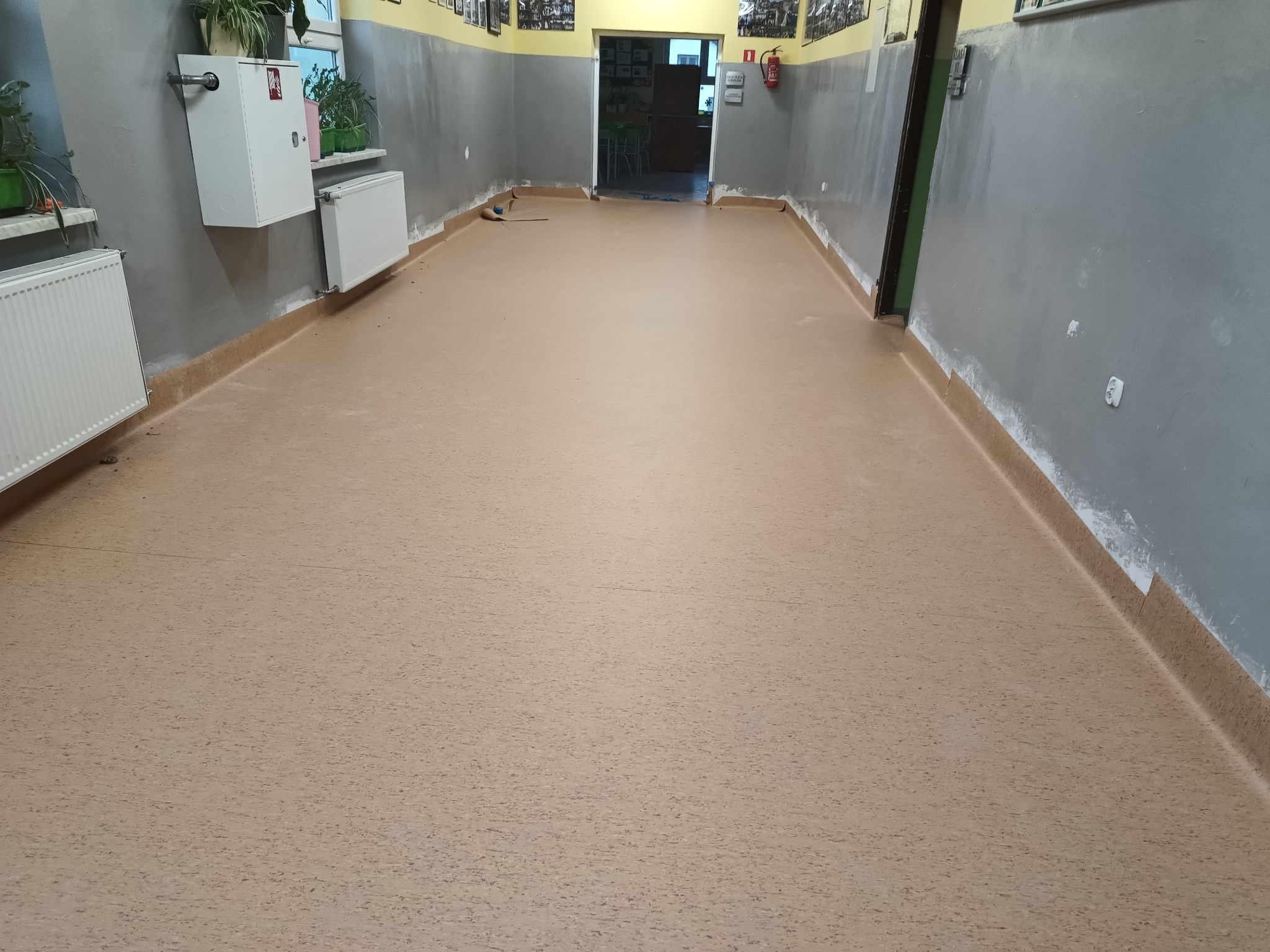 Beżowa, jednolita posadzka w korytarzu szkolnym, widoczne ślady po remoncie na ścianach i listwach przypodłogowych.