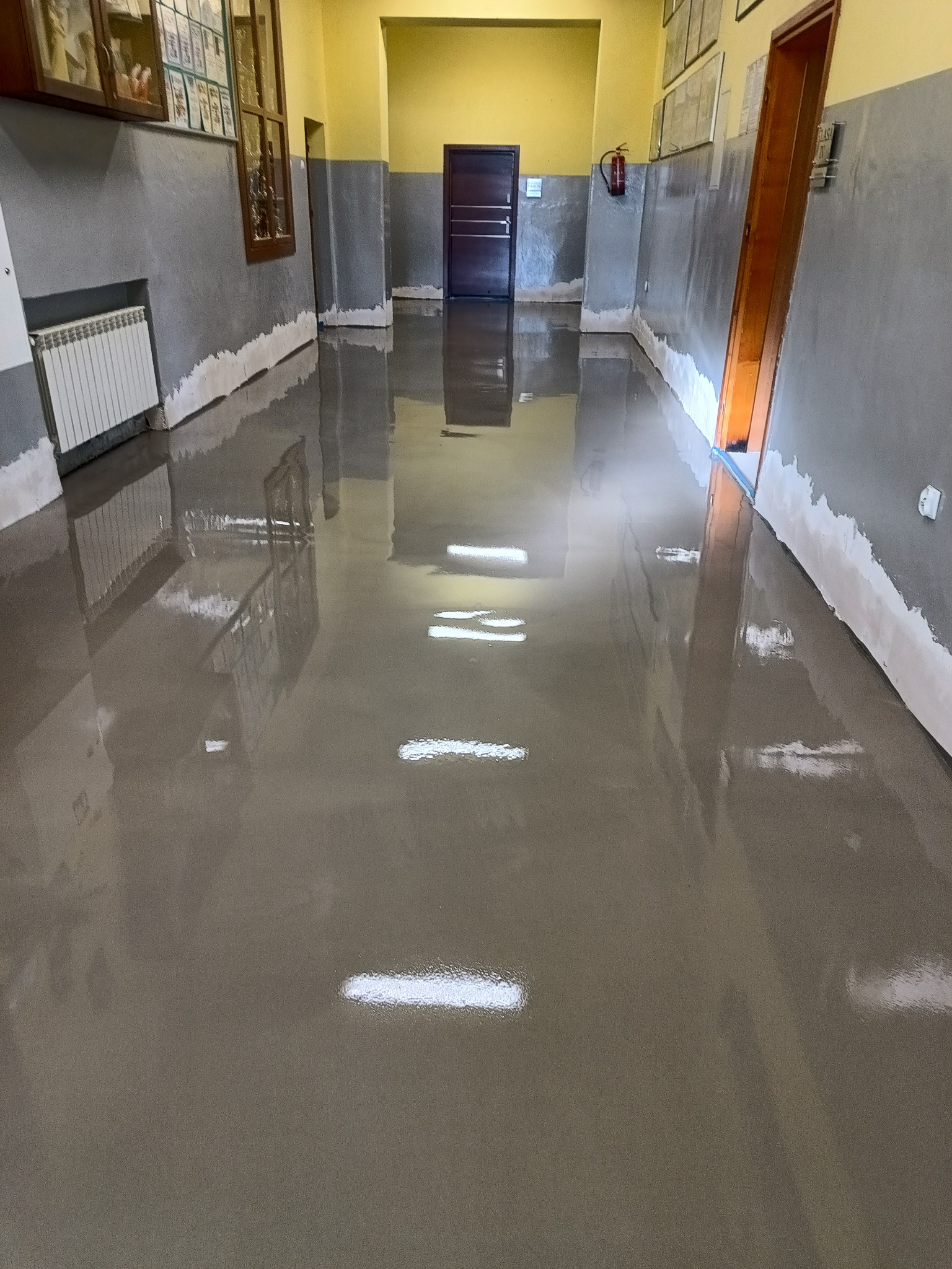 Błyszcząca, szara podłoga w korytarzu szkolnym z grzejnikiem i tablicami ogłoszeń na ścianach, odbijająca światło lamp sufitowych.