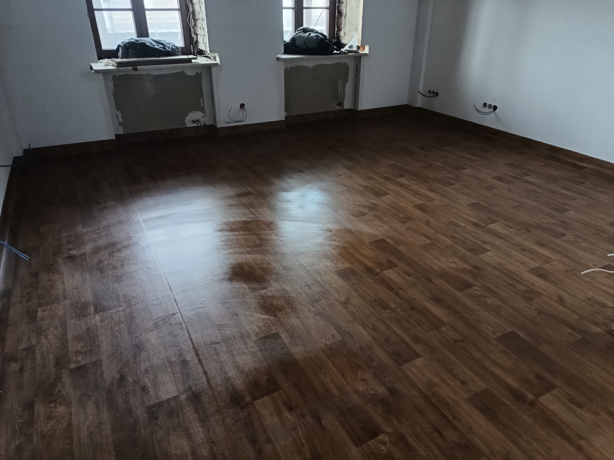 Nowo ułożona, ciemnobrązowa podłoga z paneli w pustym pokoju z dwoma oknami. Okna zabezpieczone folią. Na parapetach leżą worki z gruzem. Widoczne gniazdka elektryczne na ścianie.