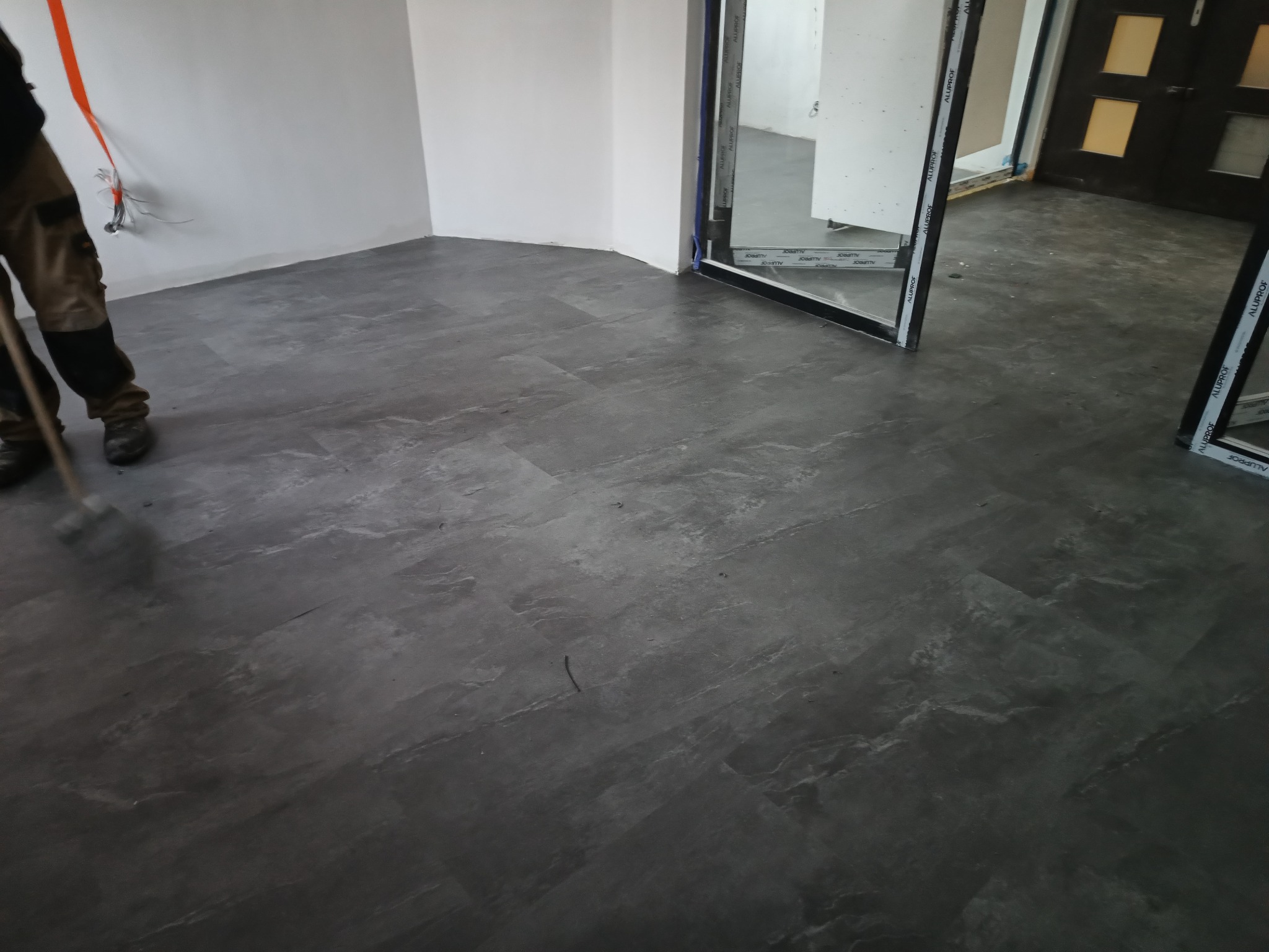 Szara podłoga w nowym pomieszczeniu z widocznymi kablami elektrycznymi wystającymi ze ściany, pracownik zamiata podłogę przed dalszymi pracami wykończeniowymi.