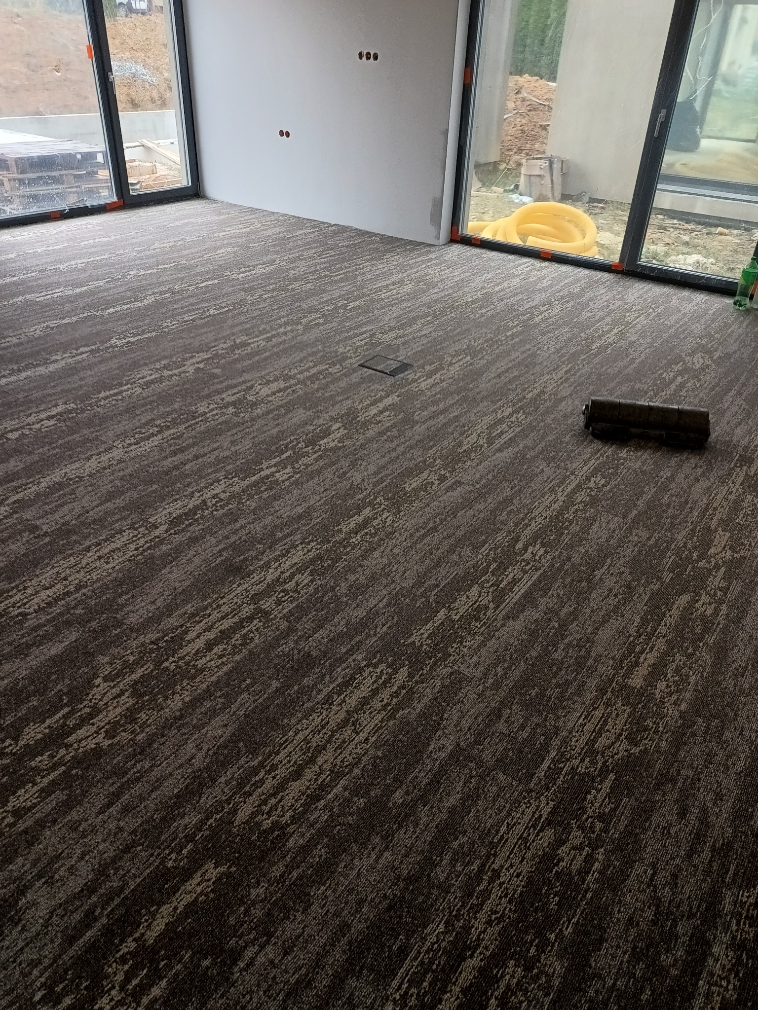 Wzorzysta, ciemna podłoga w nowym pomieszczeniu z dużymi oknami, widoczny fragment zwiniętego dywanu i żółta rura na zewnątrz.