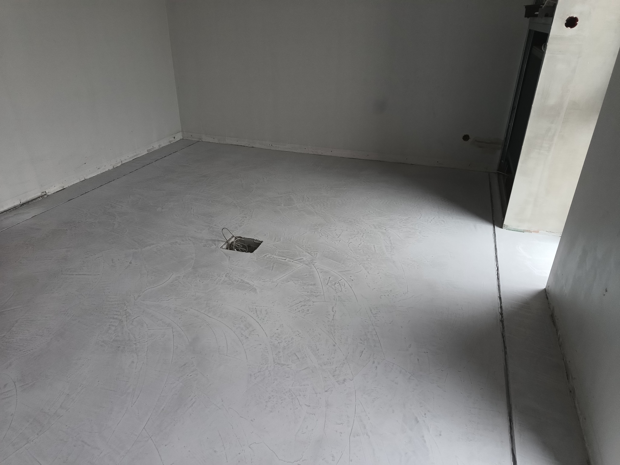 Szara, gładka powierzchnia podłogi z widocznymi śladami szpachlowania, otwór z kablami w centrum, surowe ściany w tle, widok na fragment konstrukcji ściany z prawej strony.